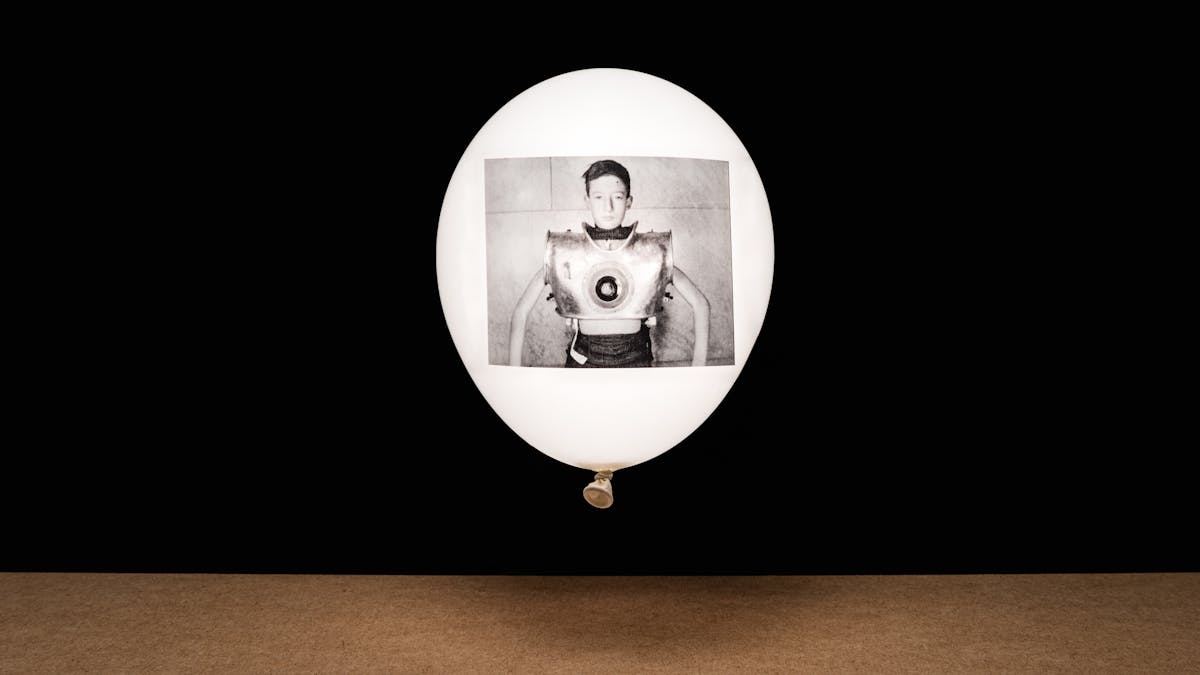 Photograph of a white inflated balloon against a black background, floating vertically above a wooden tabletop horizon line. The balloon looks like it is illuminated from within. On the side of the balloon is a rectangular, monochrome archive film still. The still shows a young boy facing the camera. His chest in encased in large metal contraption which is artificial ventilator.