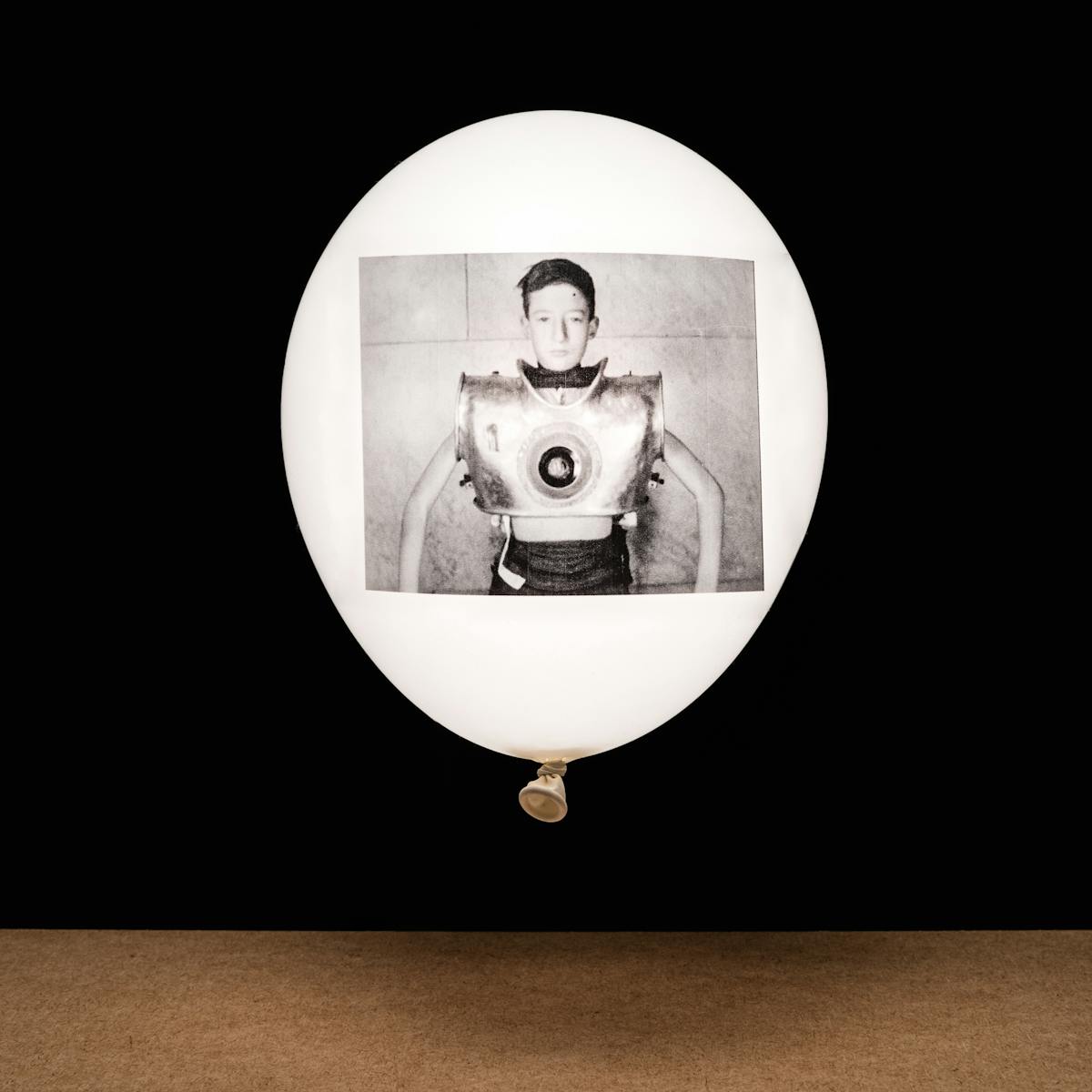Photograph of a white inflated balloon against a black background, floating vertically above a wooden tabletop horizon line. The balloon looks like it is illuminated from within. On the side of the balloon is a rectangular, monochrome archive film still. The still shows a young boy facing the camera. His chest in encased in large metal contraption which is artificial ventilator.