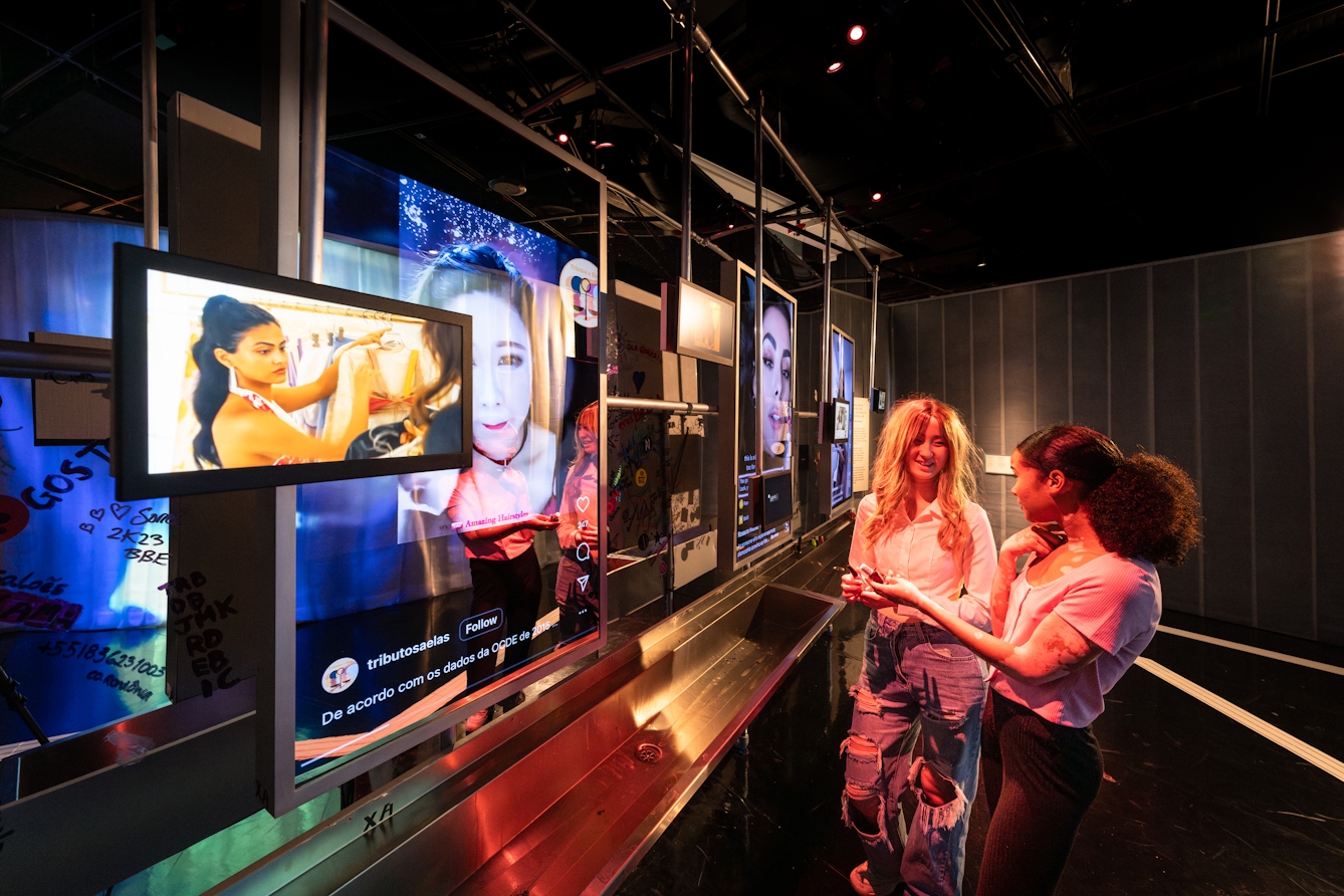 Photograph of 2 gallery visitors standing chatting by a gallery exhibit. The exhibit has a nightclub ambience and is made up of a long stainless steel sink and several different sized tv screens.