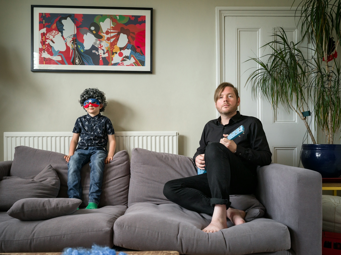 Photograph of a man and his son siting on a sofa in a living room. The son is wearing a grey curly wig and an eye mask made of a red and blue stripe and a single white star. The father is holding a small blue guitar. Behind them on the wall is a colourful poster.
