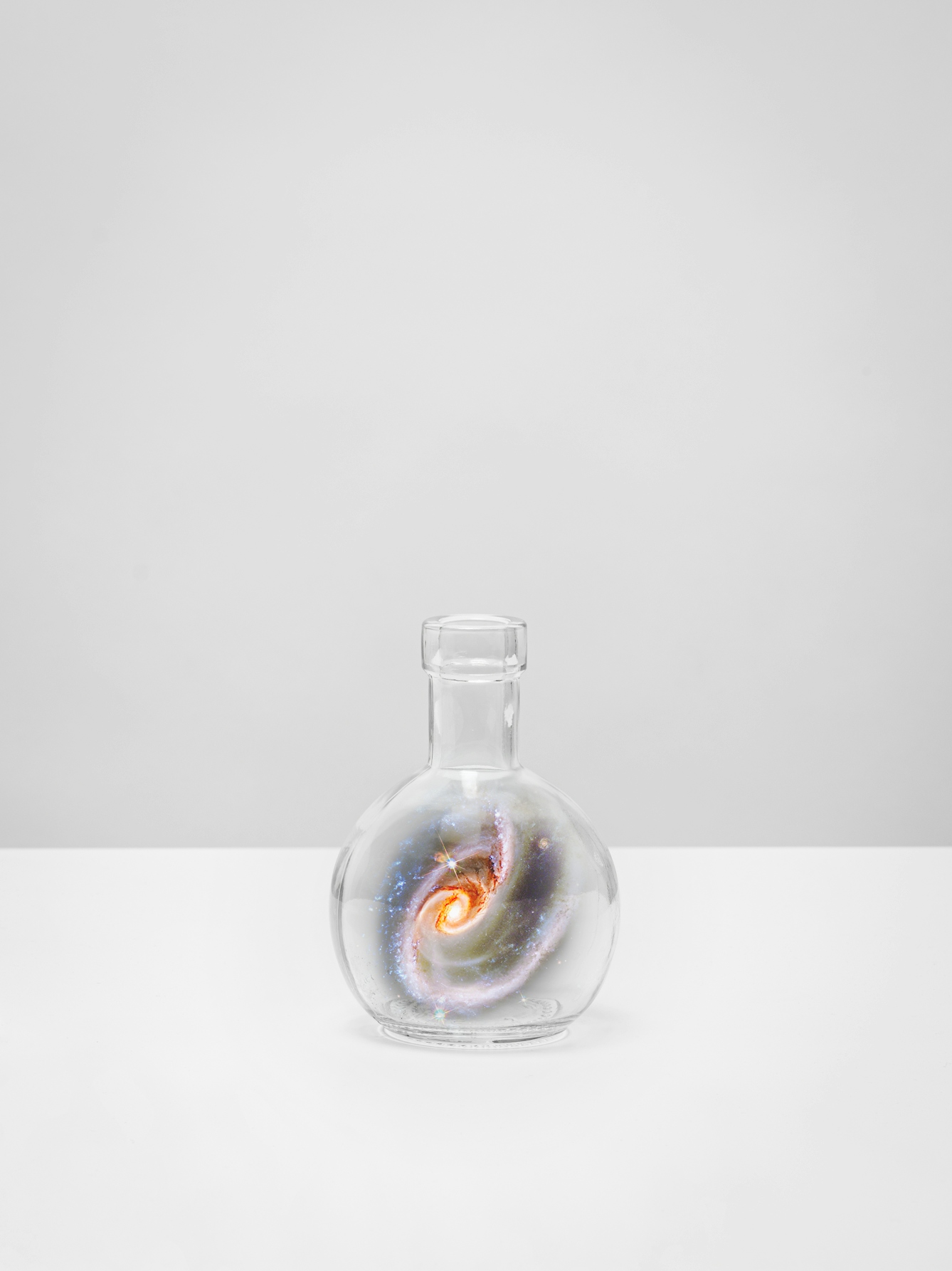Photograph of a single vertical bar, set on a white tabletop against a plain grey background. A glass flask rests on the tabletop. Swirling inside each flask is a scene of the galaxy, revealing colourful constellations, swirling stars and gaseous nebula. 