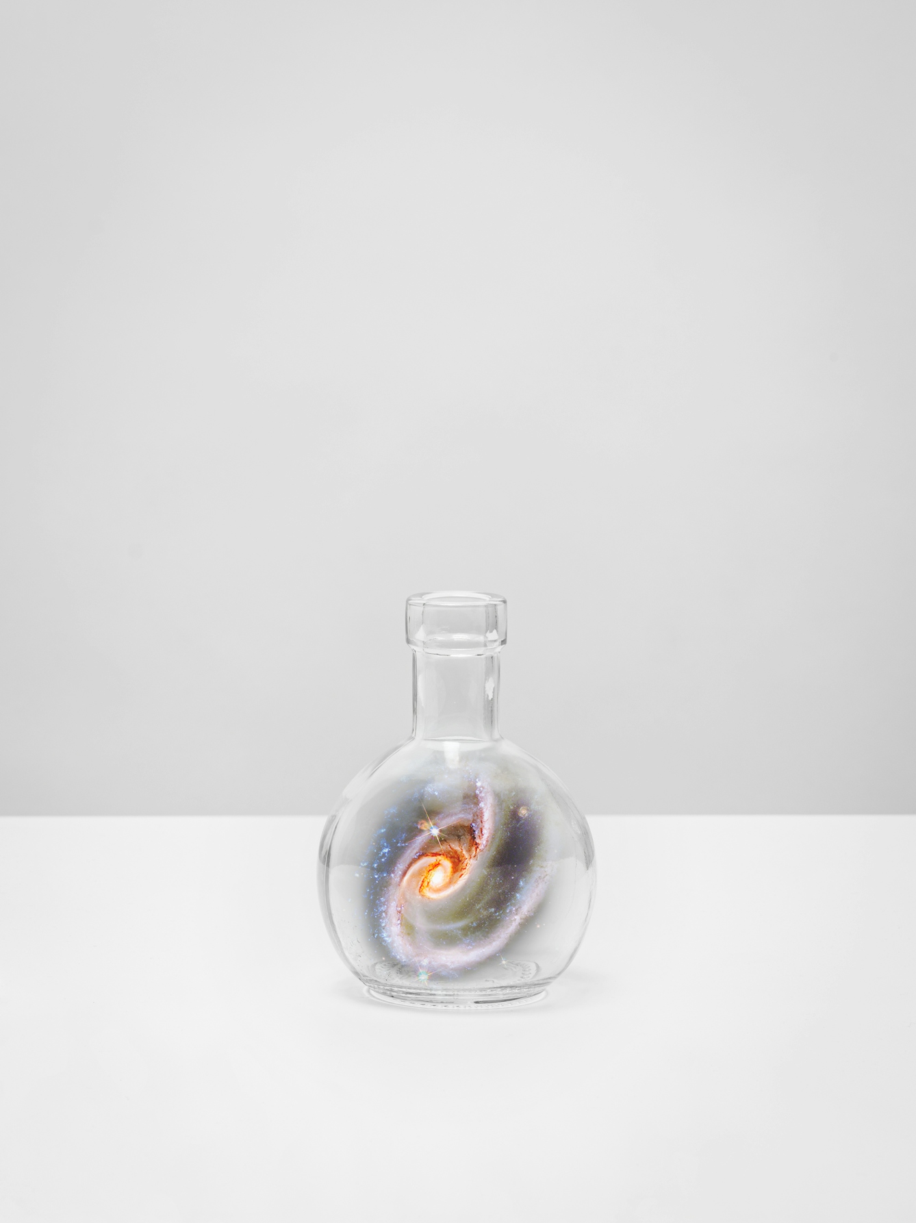 Photograph of a single vertical bar, set on a white tabletop against a plain grey background. A glass flask rests on the tabletop. Swirling inside each flask is a scene of the galaxy, revealing colourful constellations, swirling stars and gaseous nebula.