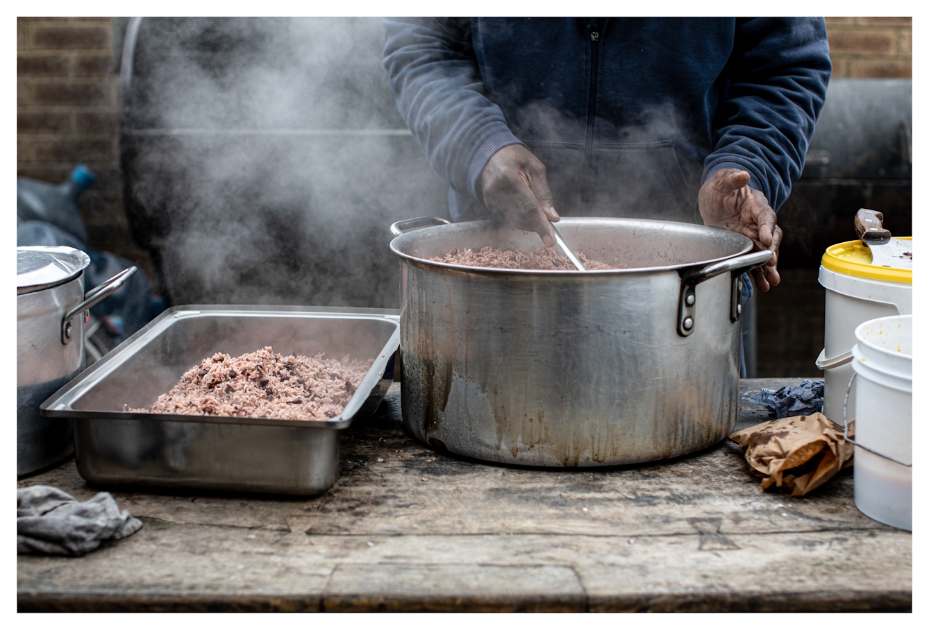 Photograph of a tabletop outside on which is a large silver cooking pot with handles full of streaming food. A person in a blue top is stood behind the table stiring the pot. Beside the large pot are other silver oven trays and saucepans.