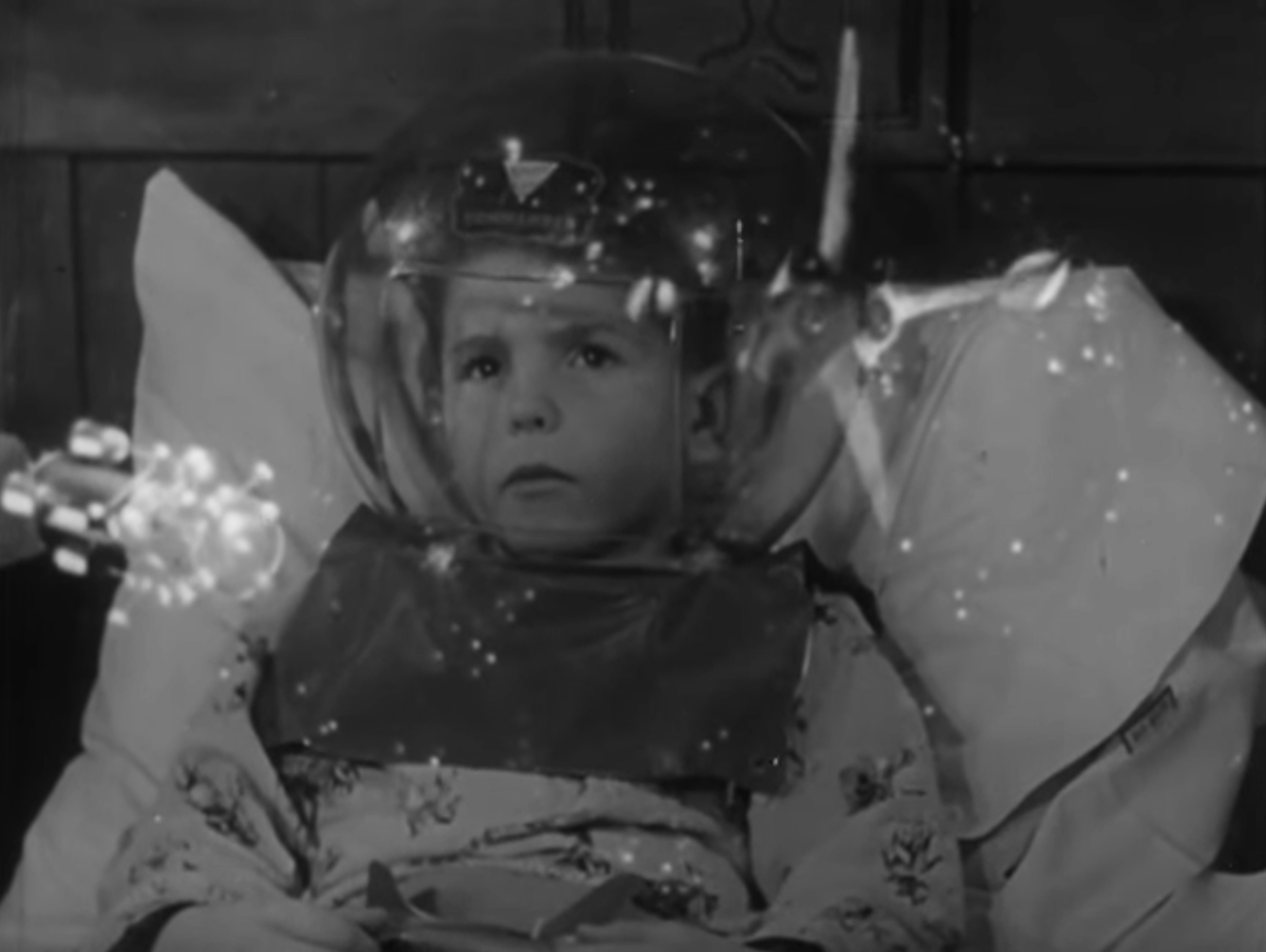 Still from black and white film featuring a boy in bed wearing a space helmet