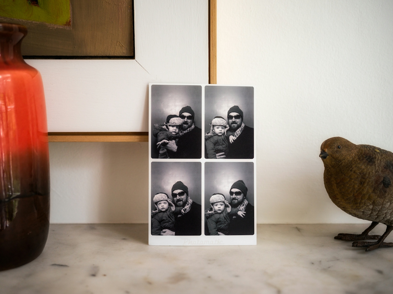 Photograph of a section of tabletop against a white wall. A set of passport four sized pictures from a photo booth are leaning against the bottom right corner of a framed picture hanging on the wall. The passport photos show a man in a hat with sunglasses holding a child who is looking directly at the camera. To the right of the print is an ornamental bird appearing into frame. To the left is a section of a red coloured ceramic vase also just appearing into frame.