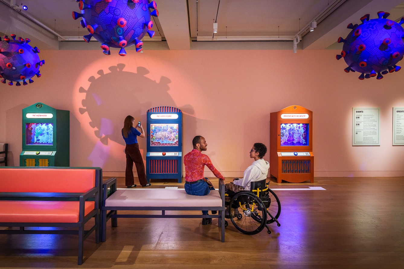 Photo of two people talking to each other in an exhibition gallery; a third person is taking a picture of an exhibit with their phone.