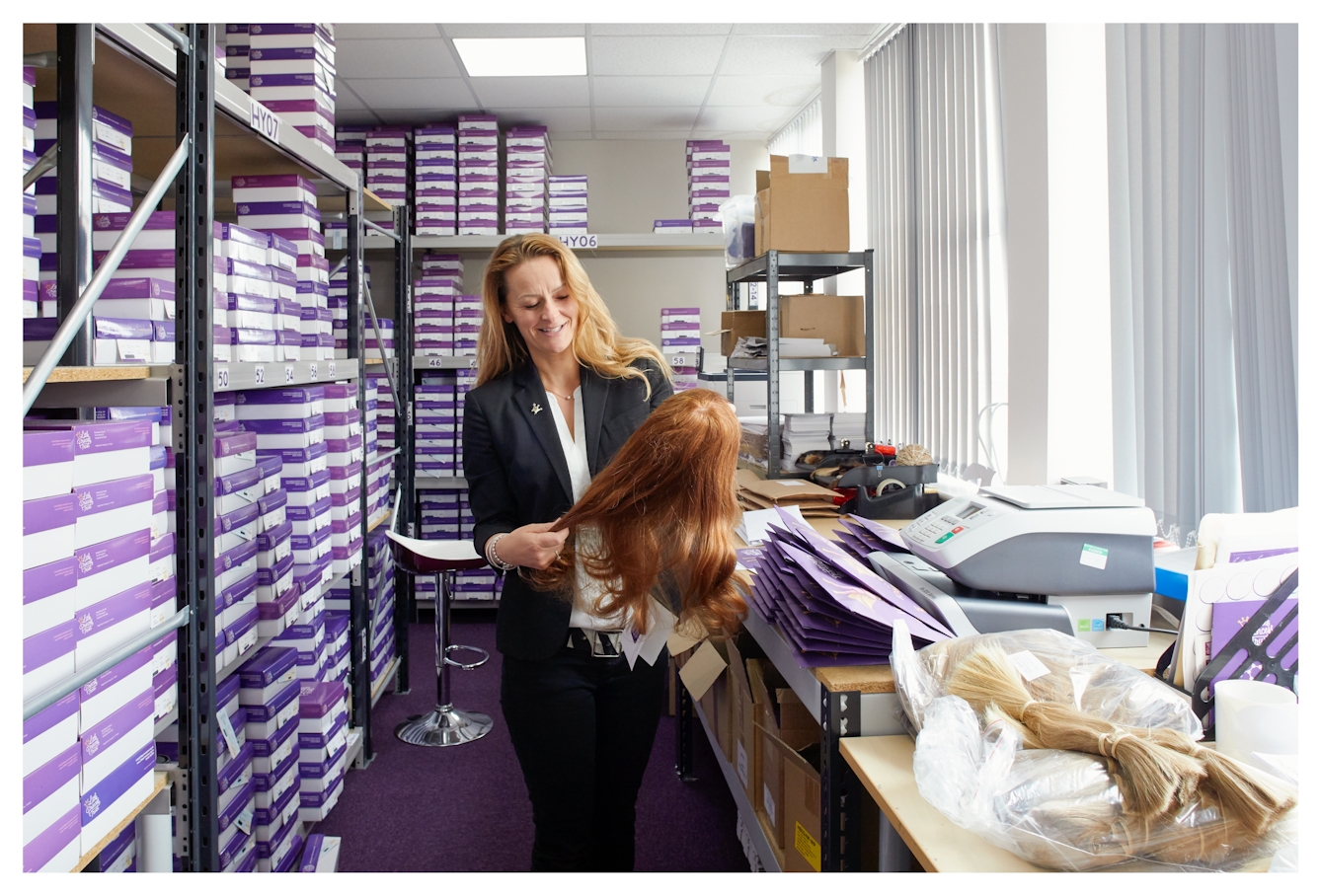 Photograph showing a woman dressed in a white blouse and dark jacket standing in a storeroom. She is looking down at her hands,  in which she is holding a ginger coloured wig. Surrounding her is shelving containing many purple and white boxes stacked on top of one another. To the right is a table top containing a piece of office equipment, purple envelopes and a bag of cut blonde hair.