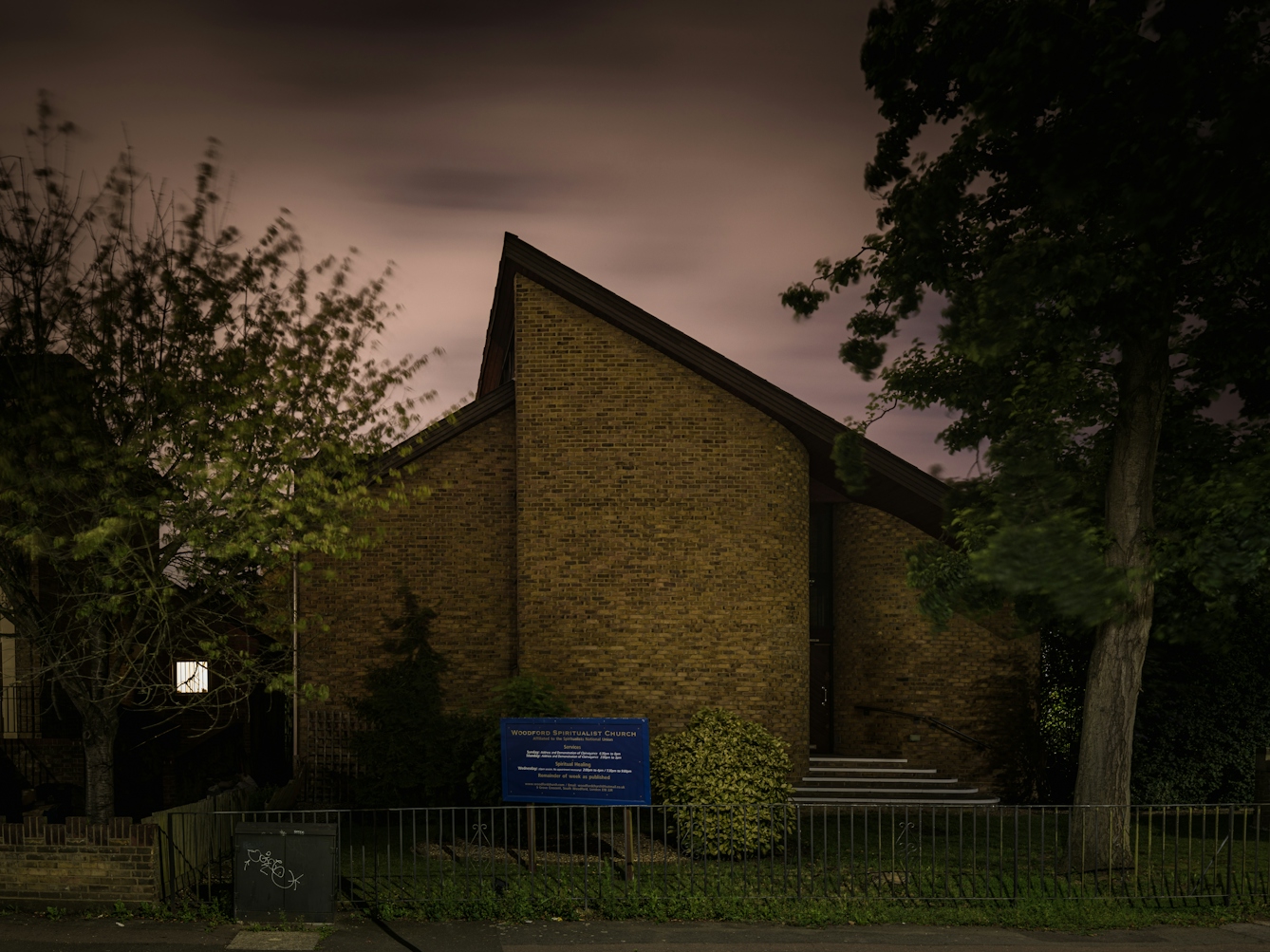 Photograph of Woodford spiritualist church, London at night.  The architecture of the church is such that it the apex of the pitched roof is off centre to the left.  The construction is bare brick with stairs to the entrance on the right.