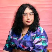 Black and white photo of a seated woman from the wait up. She has glasses and long dark, curly hair. She is smiling and looking directly at the camera.
