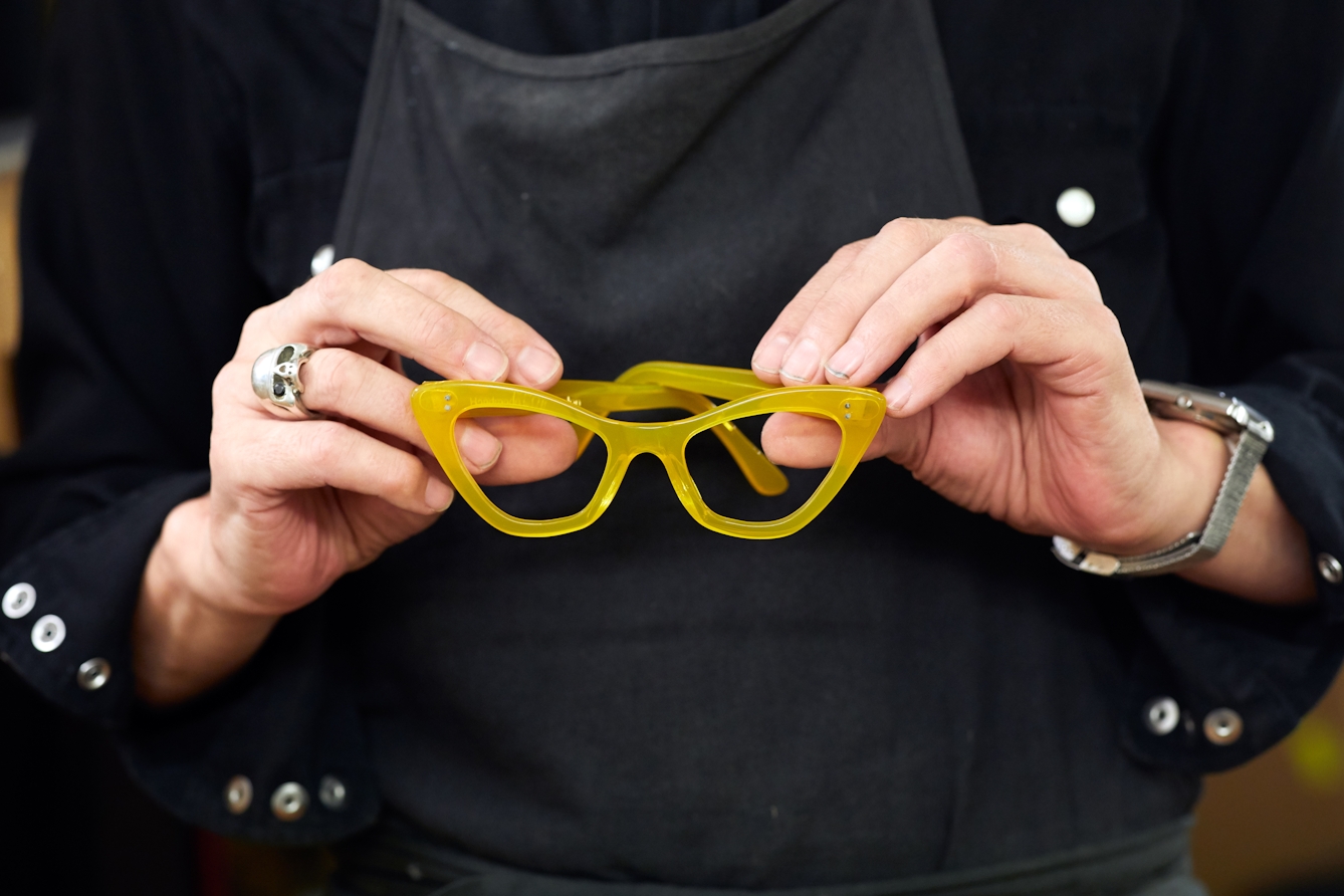 Photograph of a man wearing a black shirt and apron holding a pair of yellow framed spectacles in his hands.