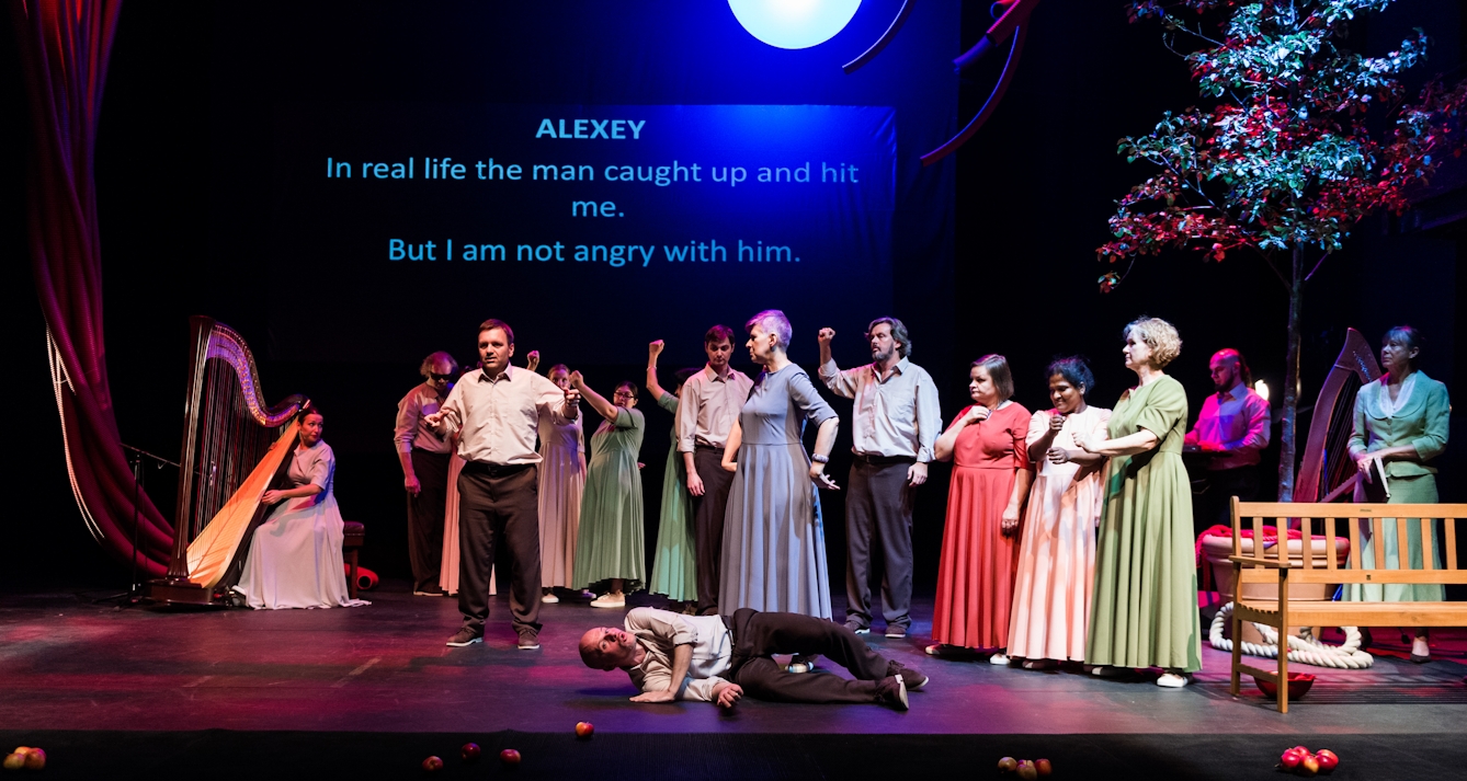 Photograph of a theatre stage. A number of actors are standing in a line in colourful formal costumes overlooking a lone male actor lying on the floor. Another male actor is delivering a soliloquy to stage centre-left. In the background to the left of the frame a female actor is playing a large floor standing harp. On the wall at the back of the set, a large projection reads “ Alexey: In real life the man caught up and hit me. But I am not angry with him”.