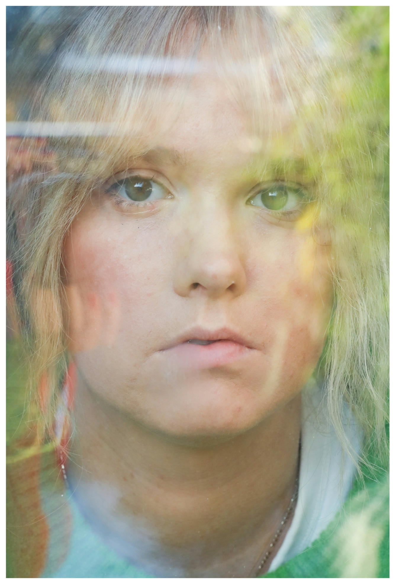 Photograph of the face and neckline of a young woman in a green jumper looking out from behind the glass of a window. Her face and shoulders are disrupted and fragmented by the reflection on the glass of the outside space which shows out-of-focus green leaves and trees. She is looking straight to camera with a direct but neutral expression.