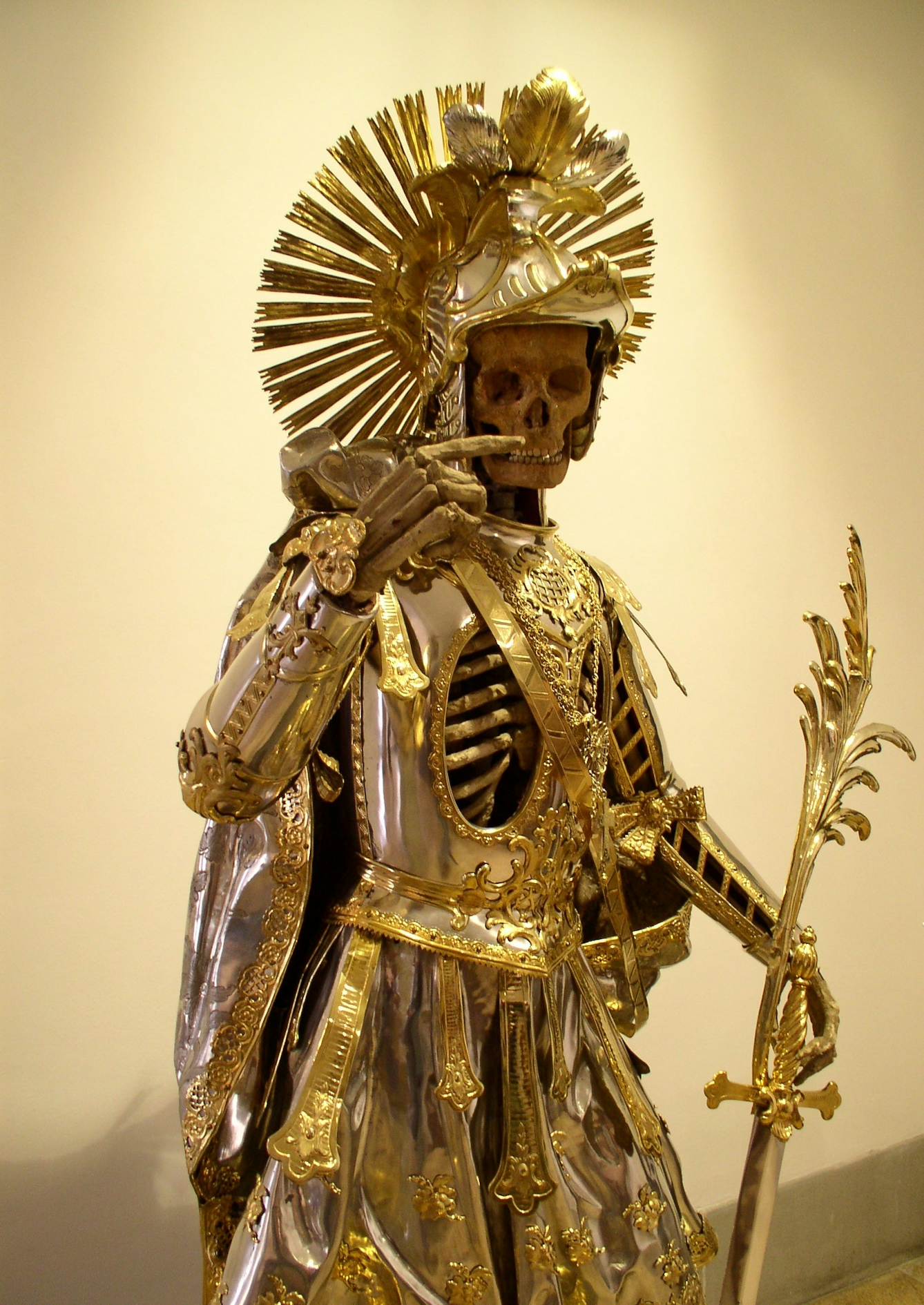 Colour photograph of a relic from the Holy Catacombs of Pancratius, showing a skeleton in intricate and lavish gold armour, with right arm raised and index finger pointing. The armour features a large helmet headdress and a highly ornate sword leaning on the left hand. 