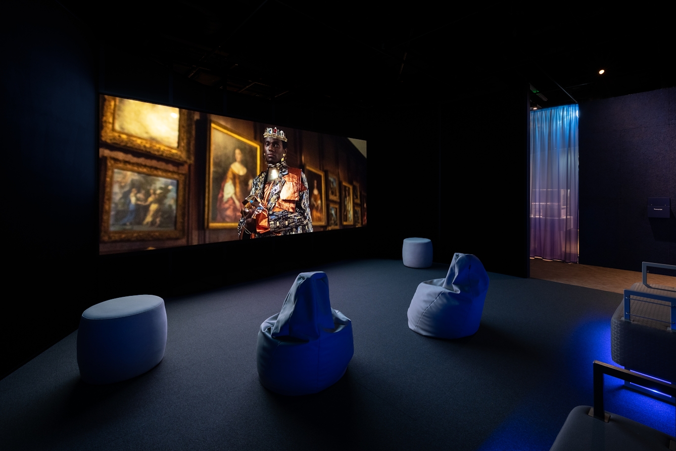 Photograph of a small room within an exhibition with a large film projected onto one wall. The still from the film shows a Black individual wearing a crown and regal gown, stood in front of a gallery of oil painted portraits. The room is furnished with stools, bean bags and benches.