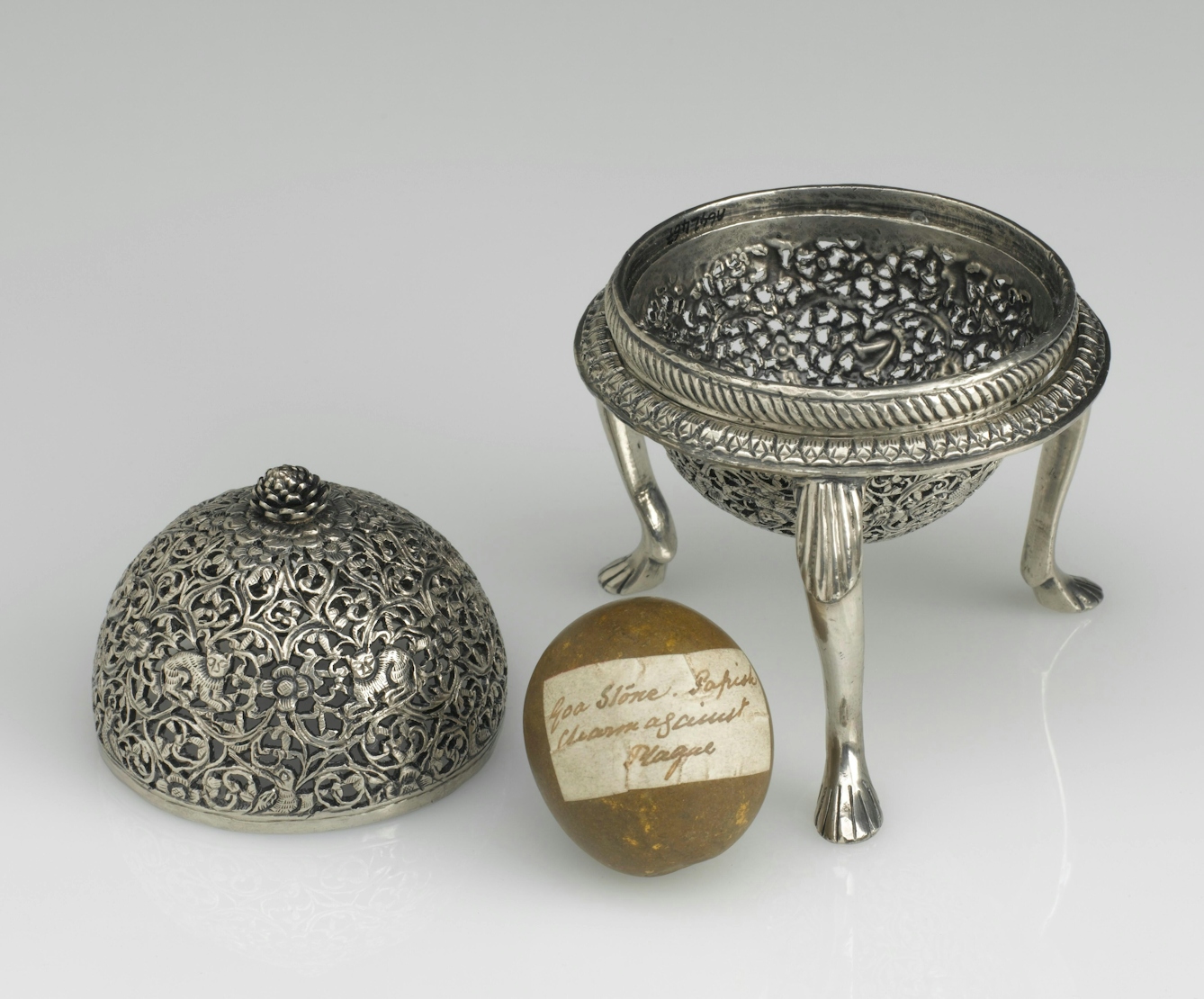 Photograph of a silver three legged case with its lid off and a goa stone with label, resting on a grey background.