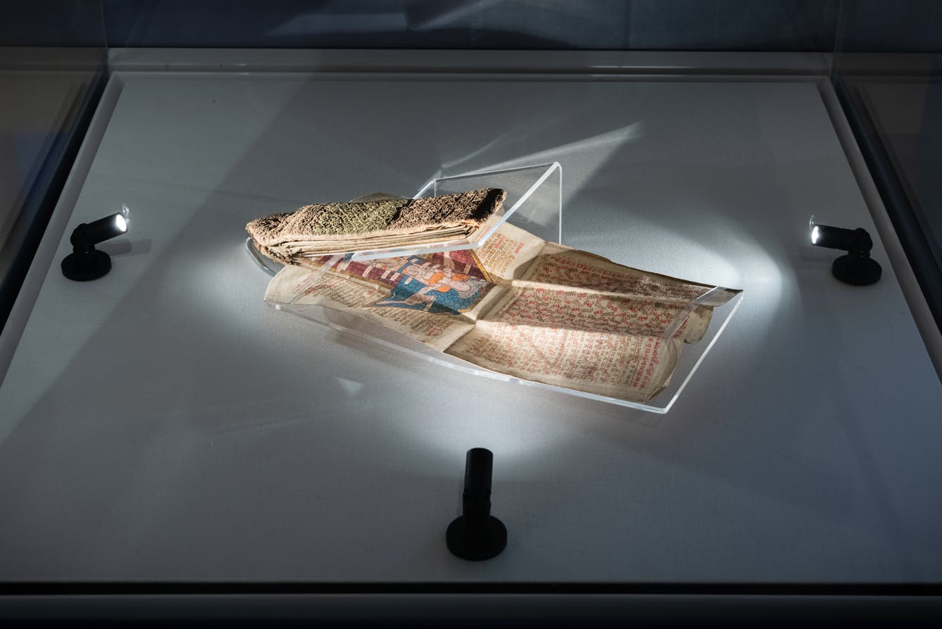 Photograph of an exhibition glass display case showing a very fragile folding almanac from the 15th century, which is partially open and supported on a perspex mount. The almanac is delicately spotlit by 3 small lights. It has a fabric textured cover and folded internal pages which are covered in hand written texts and diagrams.
