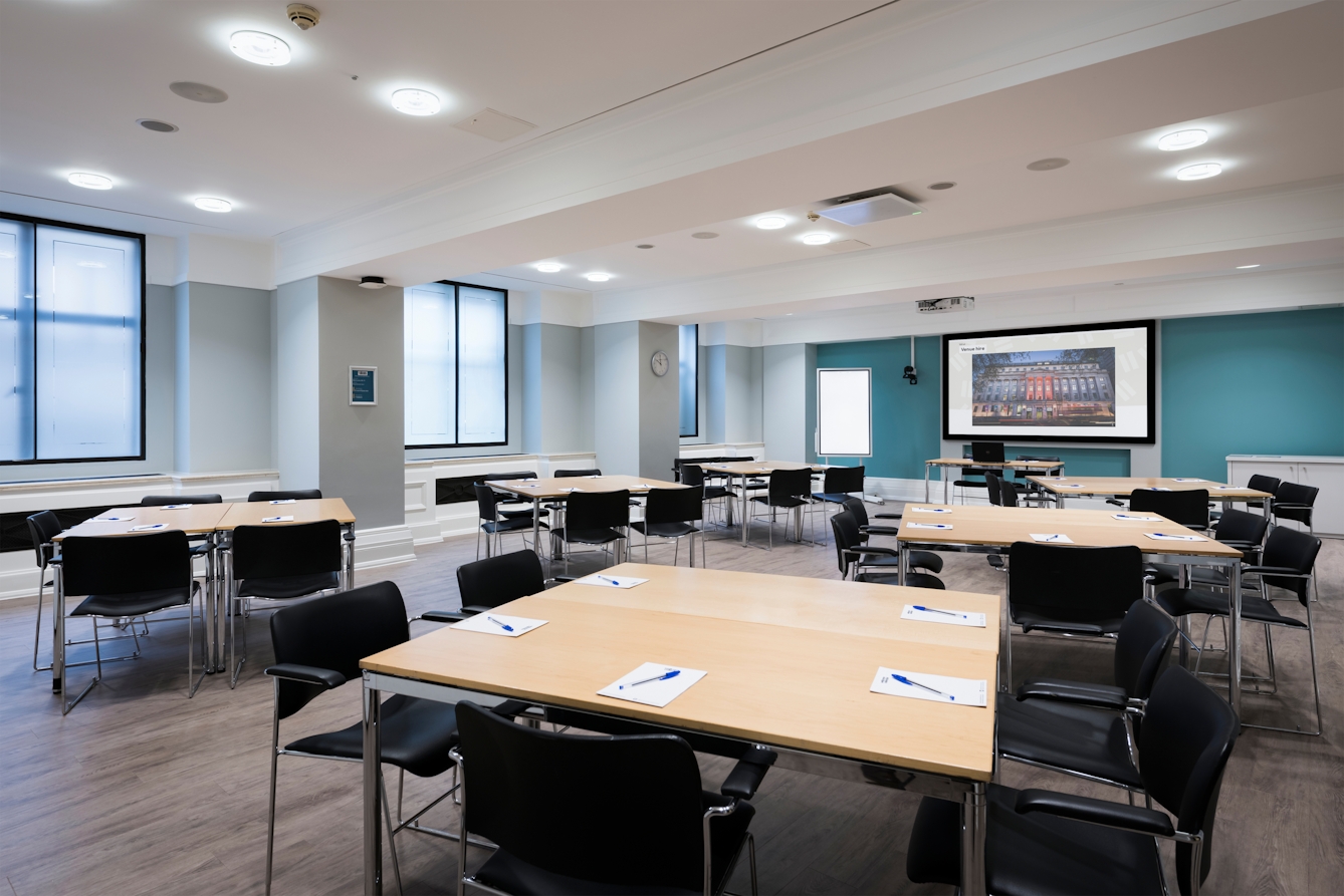 Photograph of the Franks room at the Wellcome Collection. 

Photograph shows a cabaret set-up with desks placed together in small groups. There is a projector and screen at the front of the room and several large windows.  