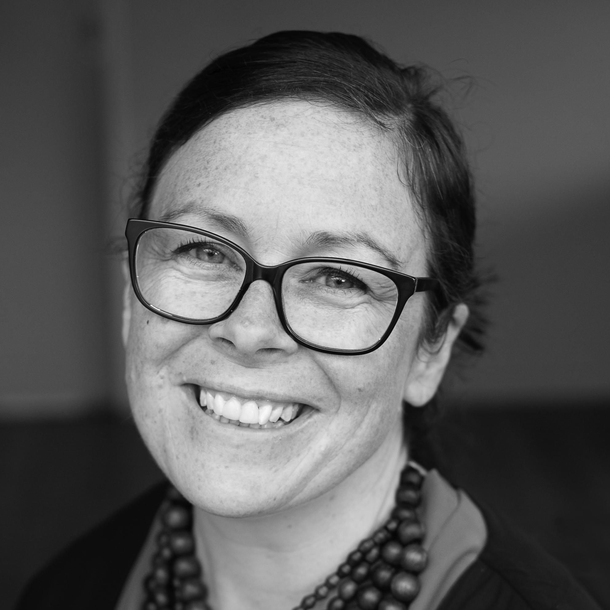 Head and shoulders photograph of a young woman smiling at the camera, wearing glasses and a beaded necklace.