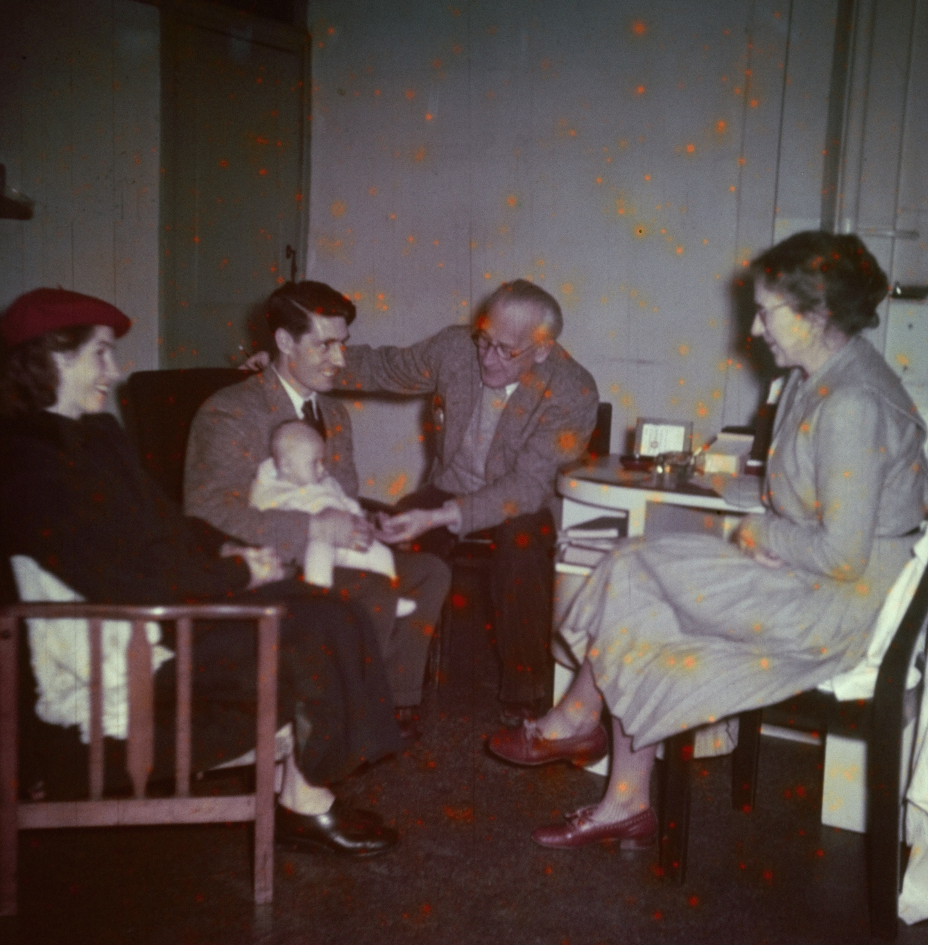 Photograph from the mid-20th Century Peckham Pioneer Centre, showing a man and woman seated on the left, the woman wearing a bright red beret and the man holding a baby on his lap. An older man reaches across towards the hand of the baby, and an older woman sits at a desk with her legs crossed, smiling at the group.