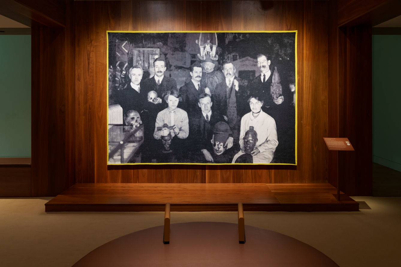Photograph of a gallery exhibition space showing large tapestry hung on a wooden wall. The tapestry is made up of a black and white, archive photograph, from the early 20th century showing a group of white men and women gathered in a group holding museum artefacts in their hands or on their laps. They are surrounded by other museum exhibits and display cases. The tapestry is framed with a bright yellow border.Photograph of a gallery exhibition space showing large tapestry hung on a wooden wall. The tapestry is made up of a black and white, archive photograph, from the late 20th century showing a group of white men and women gathered in a group within an exhibition exhibit. One of them is seated on an ornate throne with the others perched and lying around him, some holding museum artefacts in their hands. The tapestry is framed with a bright pink border. Surrounding the tapestry is a wider wooden structure containing seating and alcoves.