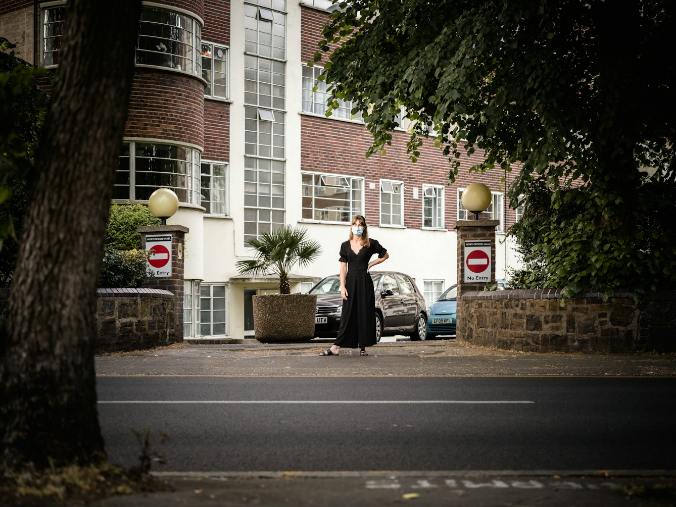 Photograph of a woman in a black dress and wearing a blue face-mask standing in the middle of an exit to a residential building. Two signs displaying the do not enter circle with white dash appear on pillars either side of the exit. The shot is captured from the other side of the road and features the trunk of a tree, the road and part of the pavement in the foreground.