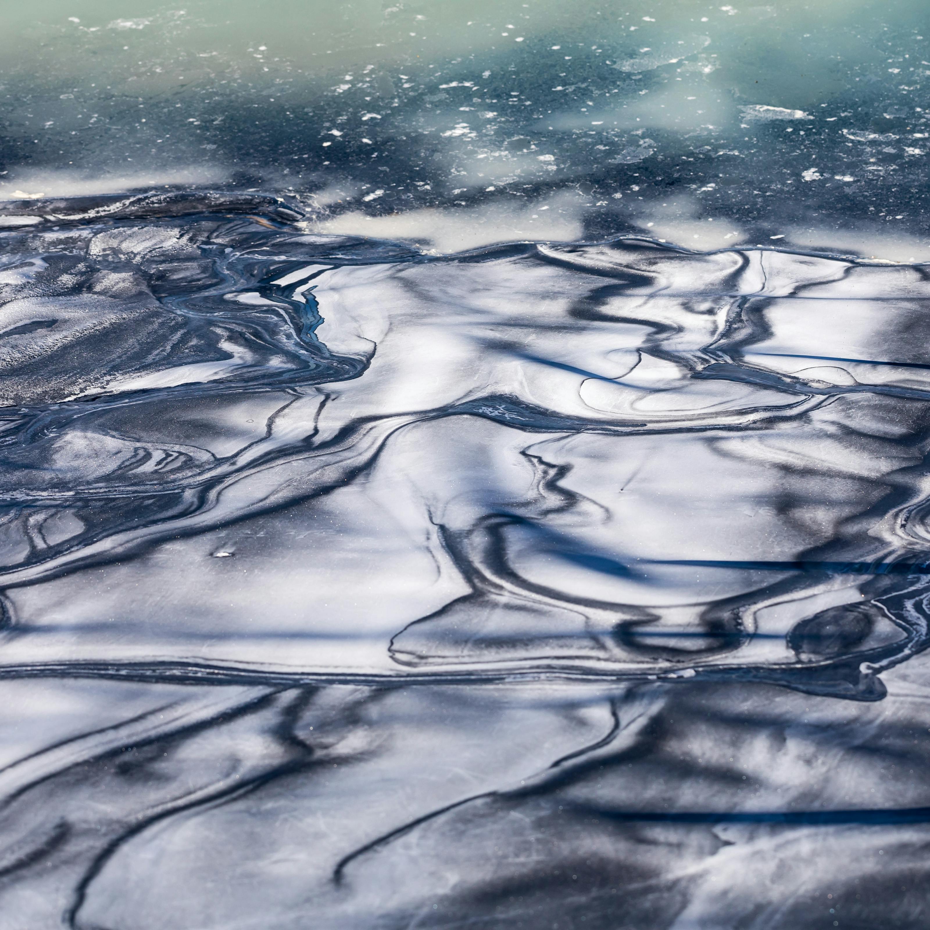 Colour photograph of salt deposits in evaporation pools. The image is abstract in appearance with few recognisable reference points. This makes it difficult to gauge the scale of the subject matter. The image itself is made up of blue, green and white tones. Most of the image seems to have a ripple pattern of whites and blues which resemble the surface of water, but which could be solid. Towards the top of the image the ripple disappear to leave a more constant tone of blues and greens, speckled with white specks and shapes.