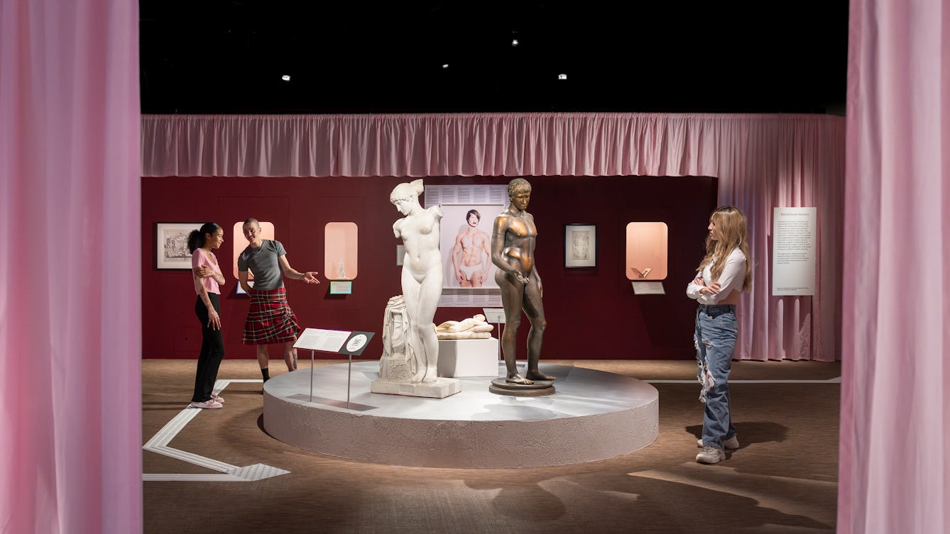 Three visitors standing in a gallery space looking at two prominent statues. A female figure in white stone on the left and a bronze male figure to the right. Between them, hung on the far gallery wall is a photographic self-portrait by the artist, Cassils.