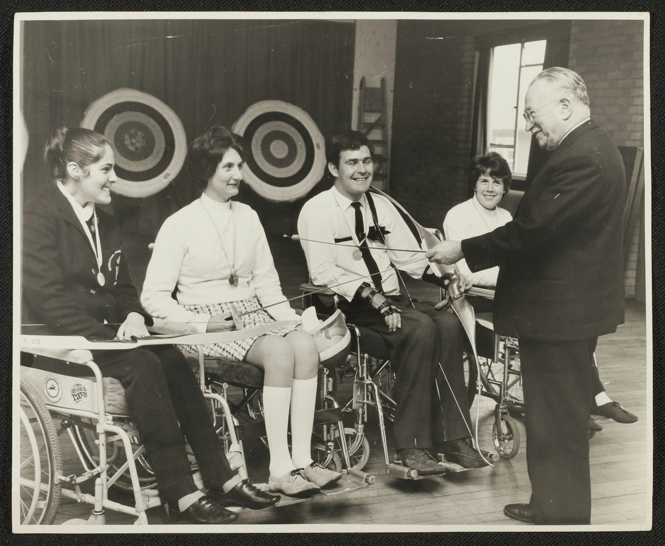 Black and white photographic print of Sir Ludwig Guttmann, on the right of the photograph, meeting with four athletes sitting in wheelchairs. Guttman wears a dark suit and shoes and faces away from the camera and toward the athletes but you can see from his profile that the bespectacled man with light hair is smiling. The athletes hold sporting equipment. The woman on the left has a beaming smile and medal and wears her hair in a bun. She holds a bow across her lap. The woman who is second from left is also smiling and has two medals around her neck. She holds a fencing foil, as does Guttmann, who appears to be speaking with her and chuckling at the moment the photograph was taken. Next is a grinning man with dark hair wearing a medal and a shirt and tie. He has straps on his wrists and holds a bow. Slightly behind Guttmann in the photograph, there is another smiling woman who also appears to have a medal ribbon around her neck. 