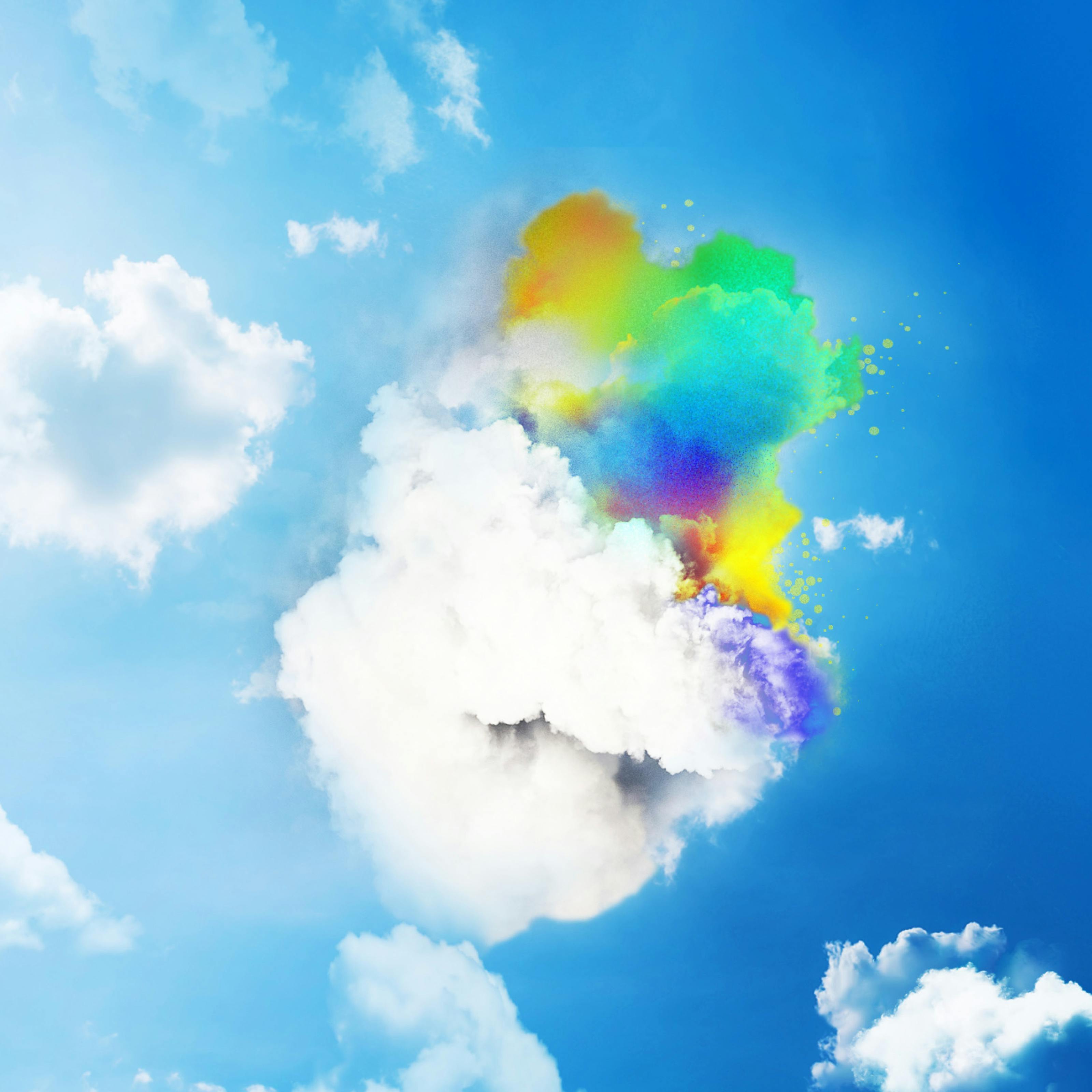 Digitally created colour image showing a bright blue sky which graduates from light blue on the left to dark blue on the right. The sky is peppered with fluffy white cloud formations. One cloud is prominently central in the frame and the top section of it has been tinted with all the colours of the rainbow, greens, reds, violets, oranges, yellows. 