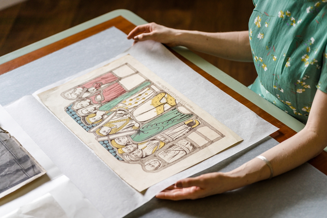 Photograph of a close-up of a woman gently holding an open archive folder in which is a large colour etching of several historical figures.