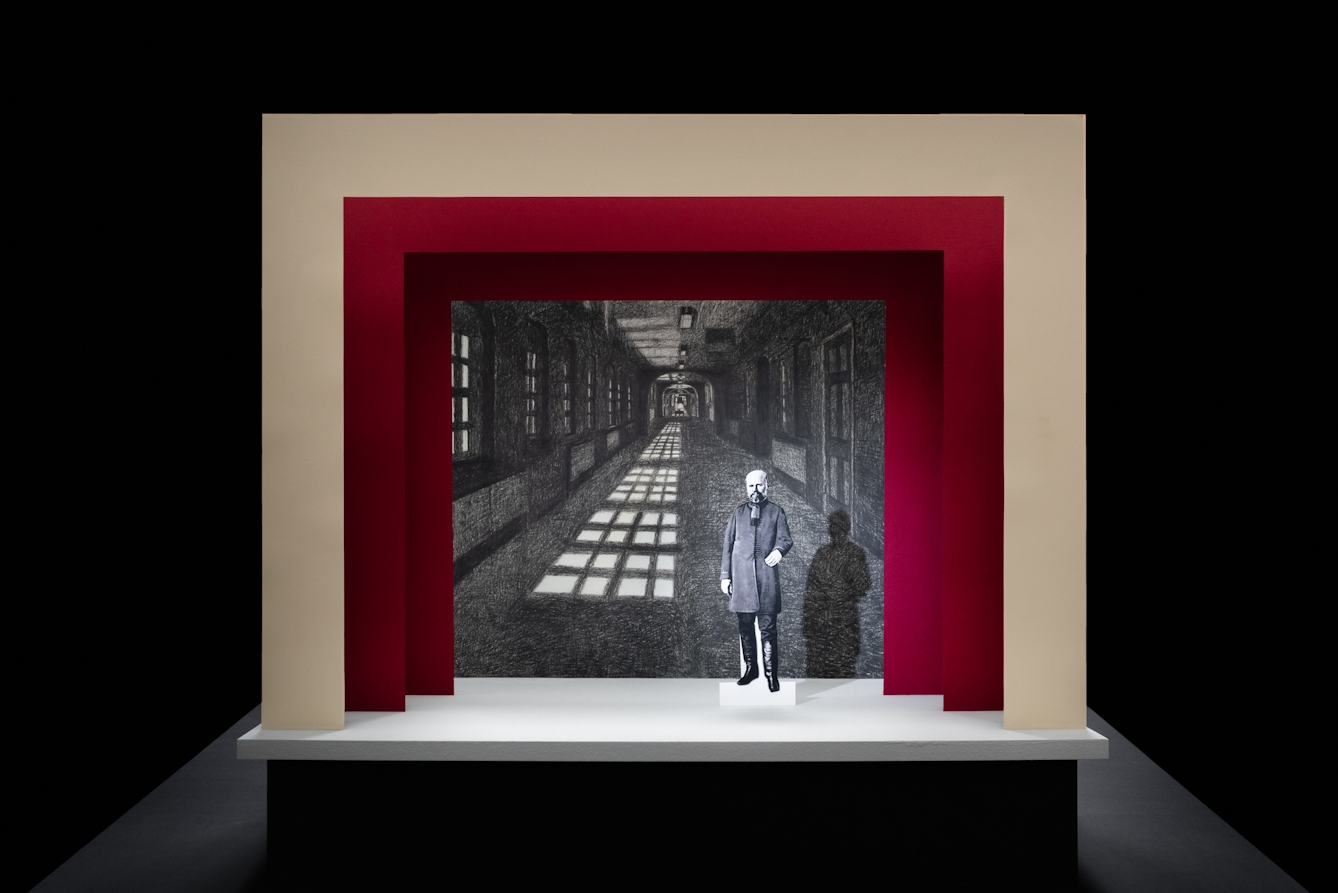 Photograph of a simple theatre stage set, made out of card. The background surrounding the stage is black. The stage floor is white and the framing of the stage is made out of 3 square edged arches, each one smaller than the other, receding backwards. The first arch is cream coloured and the other two are a red. On the stage is a small cut out illustration of a man with a beard from the early 20th century. Behind him forming the backdrop is a black and white drawing of a long corridor with windows down the left side, casting a strong shadow on the floor.