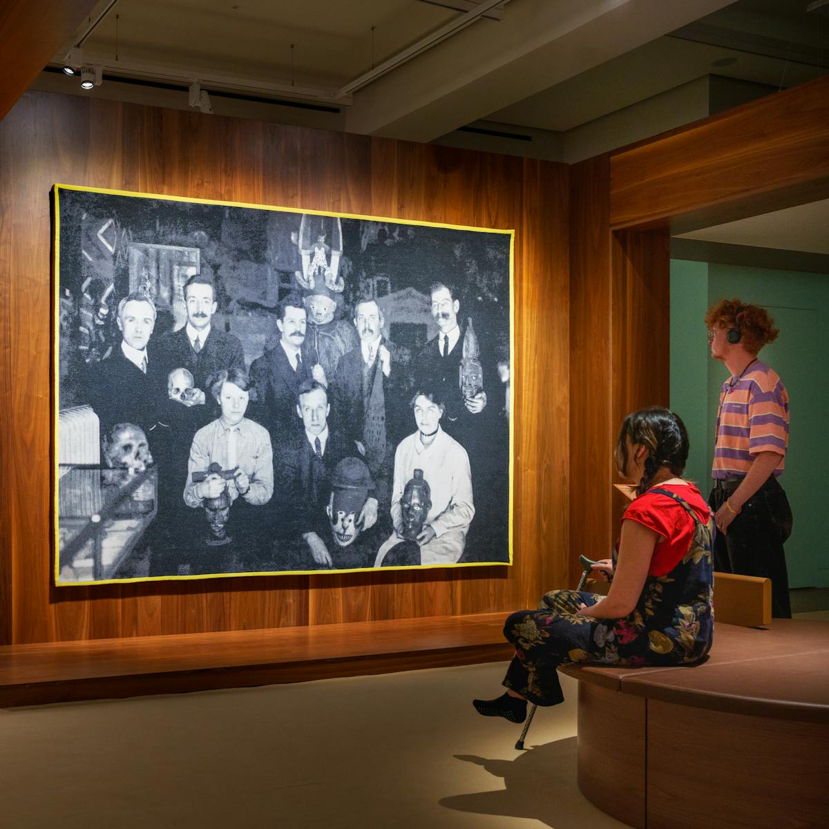 Photograph of a gallery exhibition space showing large tapestry hung on a wooden wall. The tapestry is made up of a black and white, archive photograph, from the early 20th century showing a group of white men and women gathered in a group holding museum artefacts in their hands or on their laps. They are surrounded by other museum exhibits and display cases. The tapestry is framed with a bright yellow border. Surrounding the tapestry is a wider wooden structure containing seating and alcoves. To the right of the image a gallery visitor reclines in one of the alcoves with her back to the tapestry, in contemplation. To the right two more visitors, one sitting, one standing are looking at the tapestry. The standing visitor is listening to an audio guide on headphones. The hues of the scene are warm browns and olive greens.
