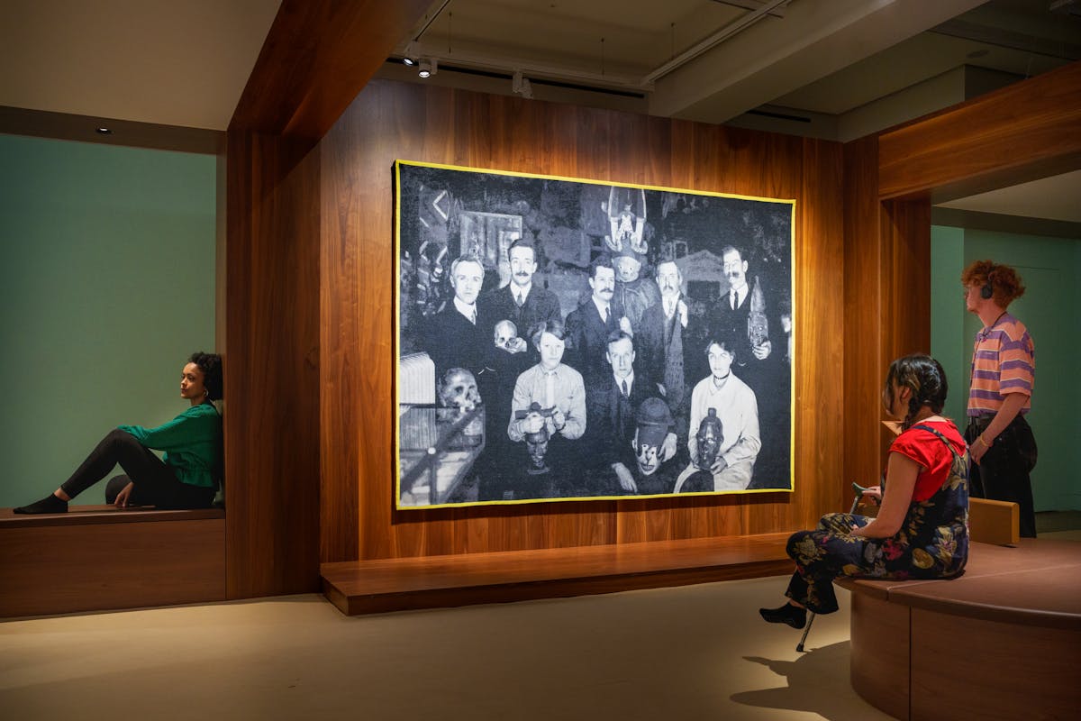 Photograph of a gallery exhibition space showing large tapestry hung on a wooden wall. The tapestry is made up of a black and white, archive photograph, from the early 20th century showing a group of white men and women gathered in a group holding museum artefacts in their hands or on their laps. They are surrounded by other museum exhibits and display cases. The tapestry is framed with a bright yellow border. Surrounding the tapestry is a wider wooden structure containing seating and alcoves. To the right of the image a gallery visitor reclines in one of the alcoves with her back to the tapestry, in contemplation. To the right two more visitors, one sitting, one standing are looking at the tapestry. The standing visitor is listening to an audio guide on headphones. The hues of the scene are warm browns and olive greens.