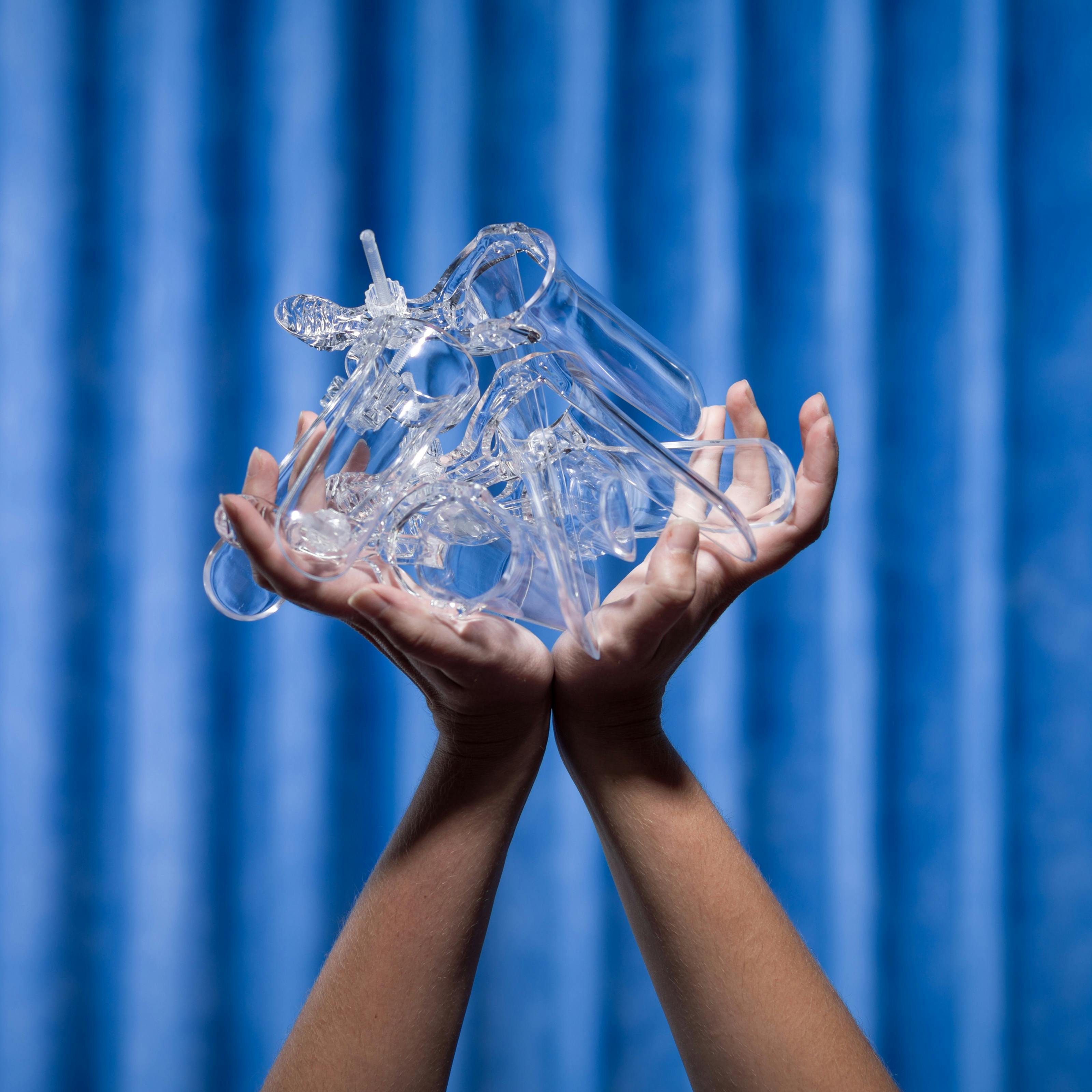 Photograph of two hands struggling to hold several vaginal speculums shot against the background of blue antibacterial clinical curtains.