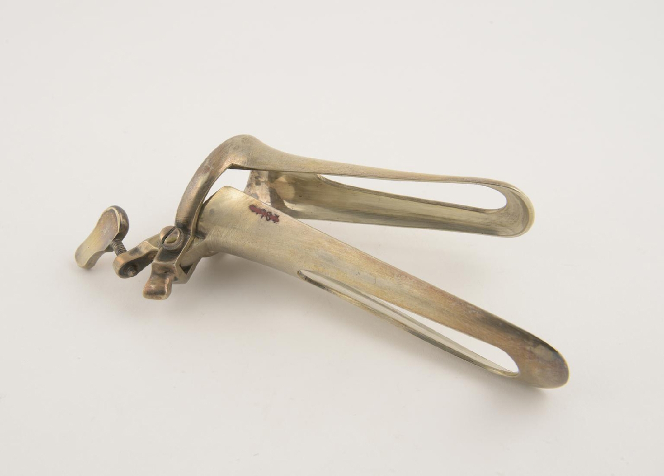 The cusco vaginal speculum, late 19th century. A silvery metal instrument consisting of two long curved parts that open and close like a duck's bill. The parts and connected at the top to a circular ring that moves them via a small screw mechanism. 