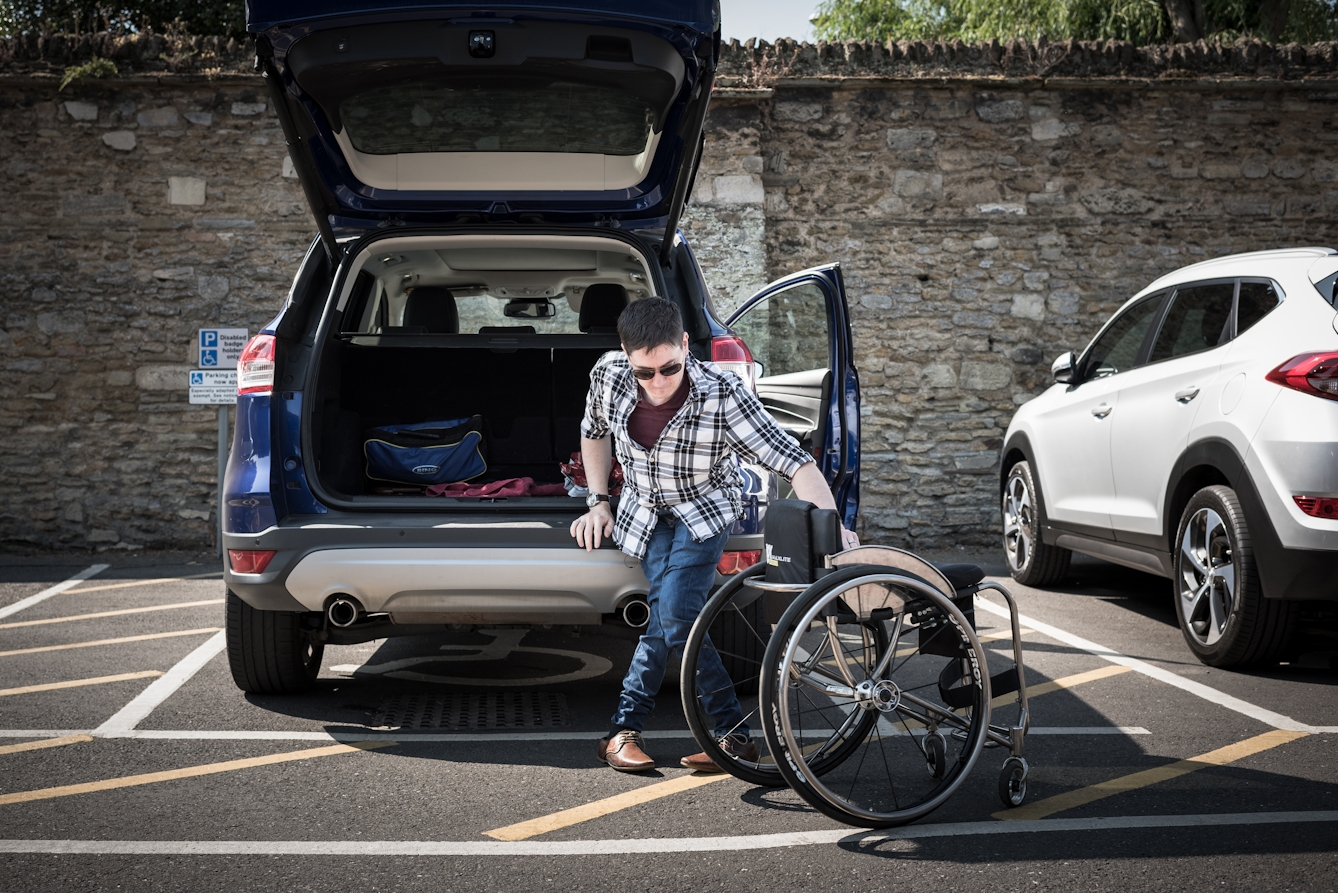 Photograph of a man unloading his wheelchair from the back of his car in a car park.