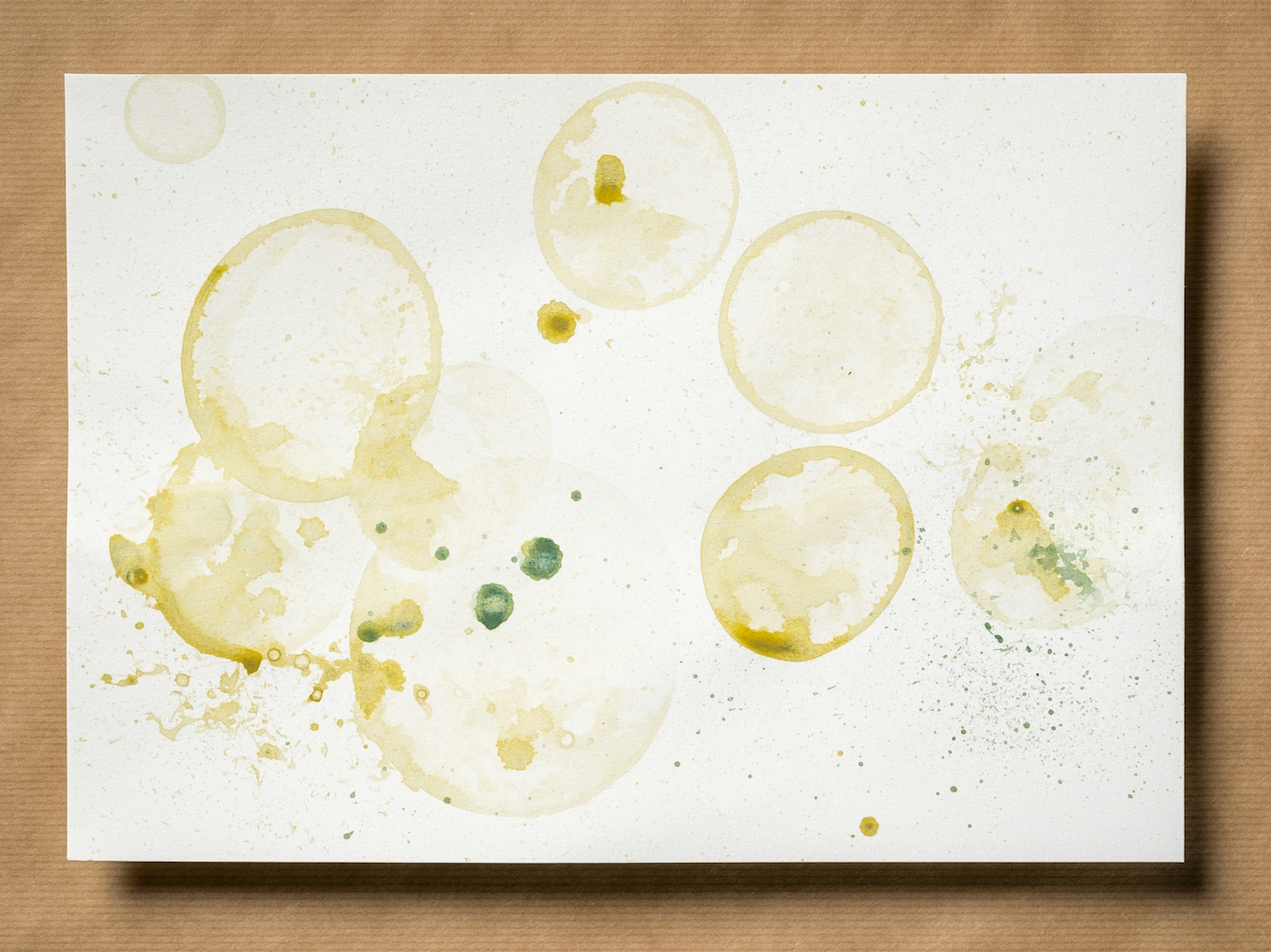 Photograph of an original artwork on watercolour paper. The artwork is resting on a brown parcel paper background. The artwork shows many varying in size droplet spatters and rings, made with coloured ink. The ink colours are dark and light greens and yellows. The spatters seem random in distribution.