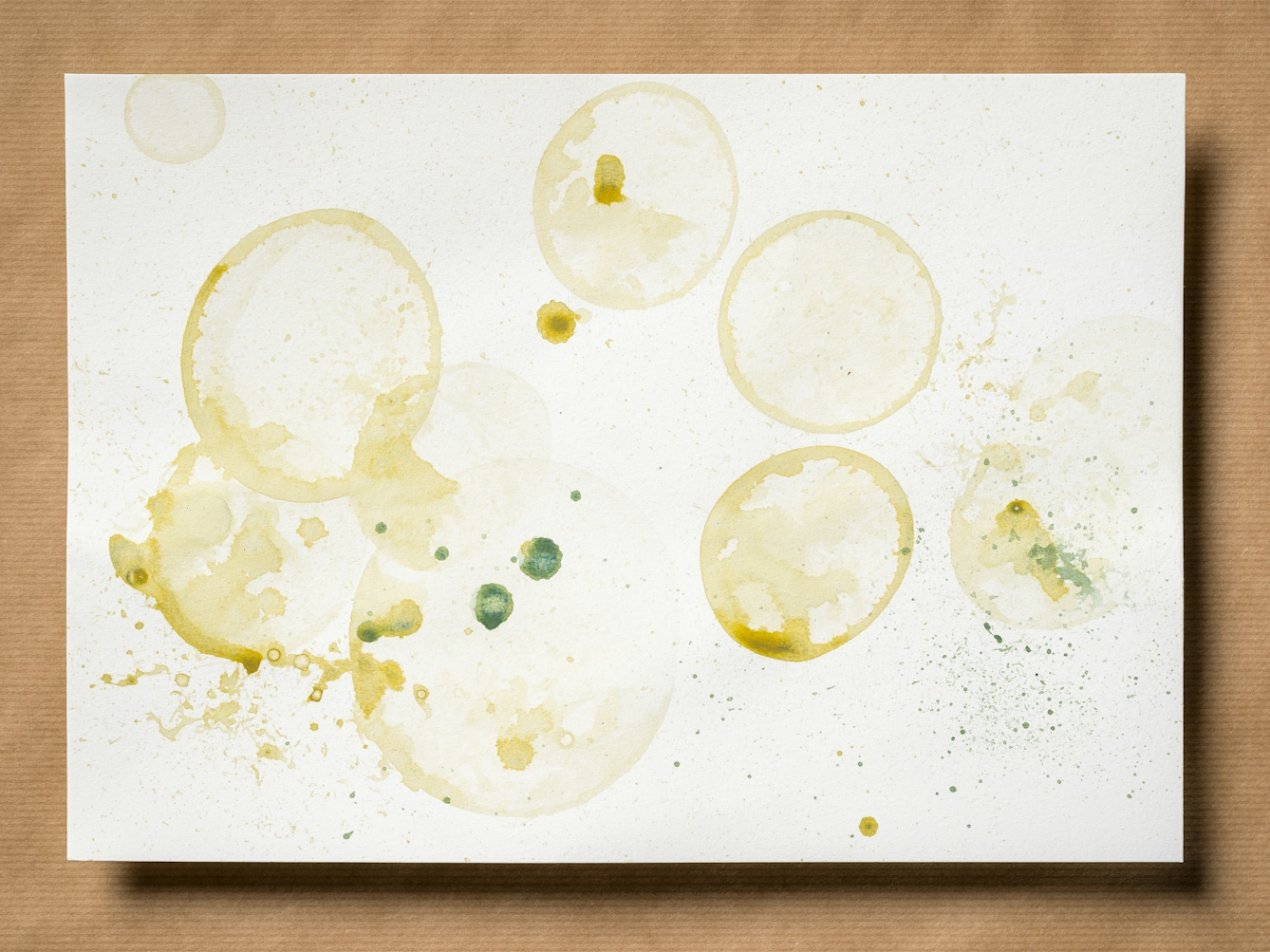 Photograph of an original artwork on watercolour paper. The artwork is resting on a brown parcel paper background. The artwork shows many varying in size droplet spatters and rings, made with coloured ink. The ink colours are dark and light greens and yellows. The spatters seem random in distribution.