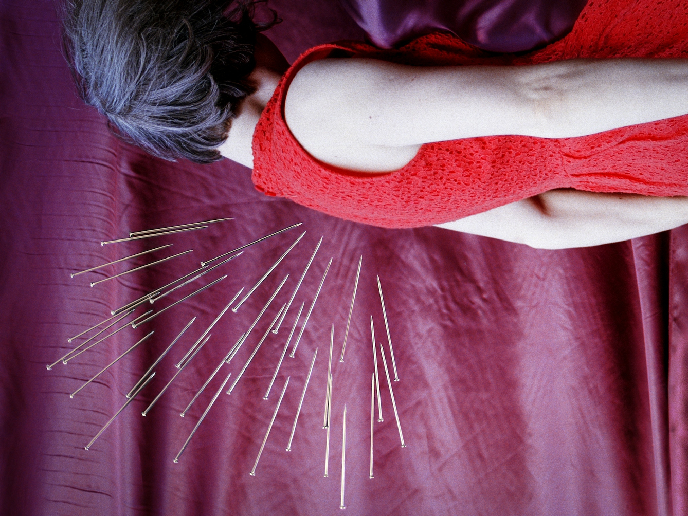 Detail from a larger artwork created with a colour photographic print of a female figure in a bright red dress, set against a purple and blue draped silk background. She is lying horizontal in the frame. Her face is obscured by her hair. Her body is surrounded by groups of dress pins, laid on top of the photographic print. The pins are arranged as if they are a flight of arrows directed at her body. One group attacks her back from underneath.