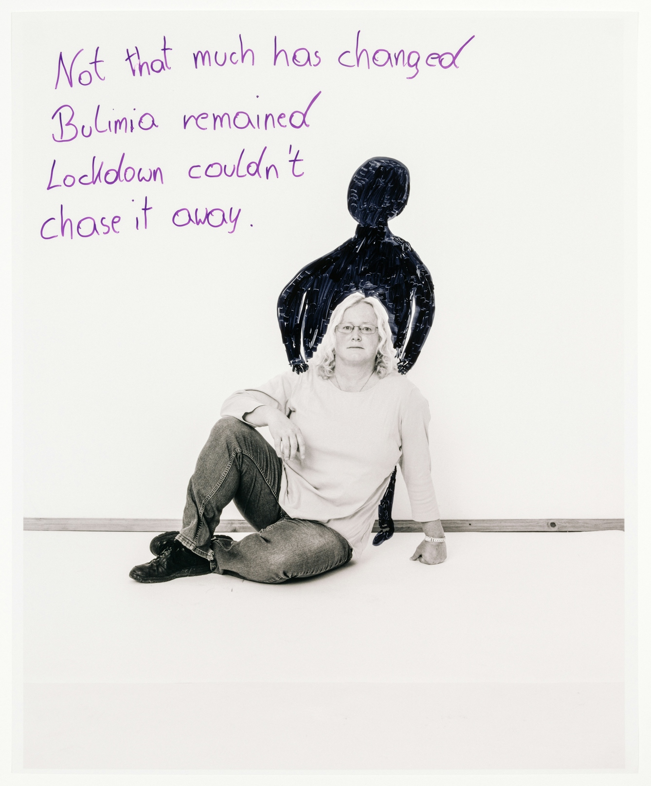 Full body photograph of a woman sitting on the floor with her legs crossed, with one arm resting on her knee and the other supporting her body upright.
Hand drawn behind her is a black figure with its hands resting on the shoulders of the woman.  Behind her, the words “Not that much has changed, Bulimia remained, Lockdown couldn’t chase it away” are written in purple marker.
