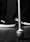 Black and white photograph of the roller tip end of a white cane resting on a wooden floor. The image is close-up on the cane so the chipped paint can bye clearly seen. In the background the feet of the person holding the cane can eb seen, white soled trainers and black trousers. The background is a dark grey making the cane a stark feature within the image.