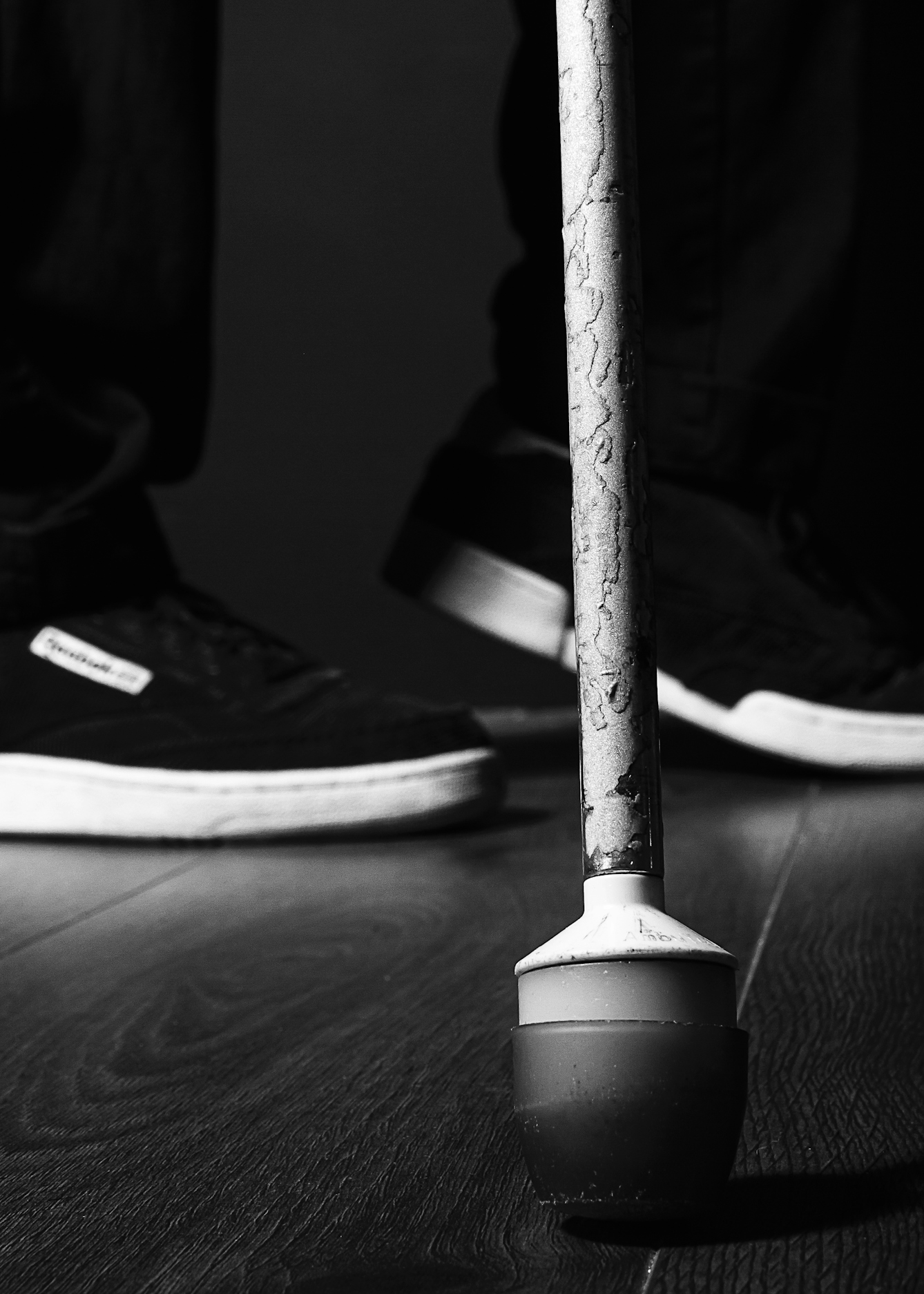 Black and white photograph of the roller tip end of a white cane resting on a wooden floor. The image is close-up on the cane so the chipped paint can bye clearly seen. In the background the feet of the person holding the cane can eb seen, white soled trainers and black trousers. The background is a dark grey making the cane a stark feature within the image.