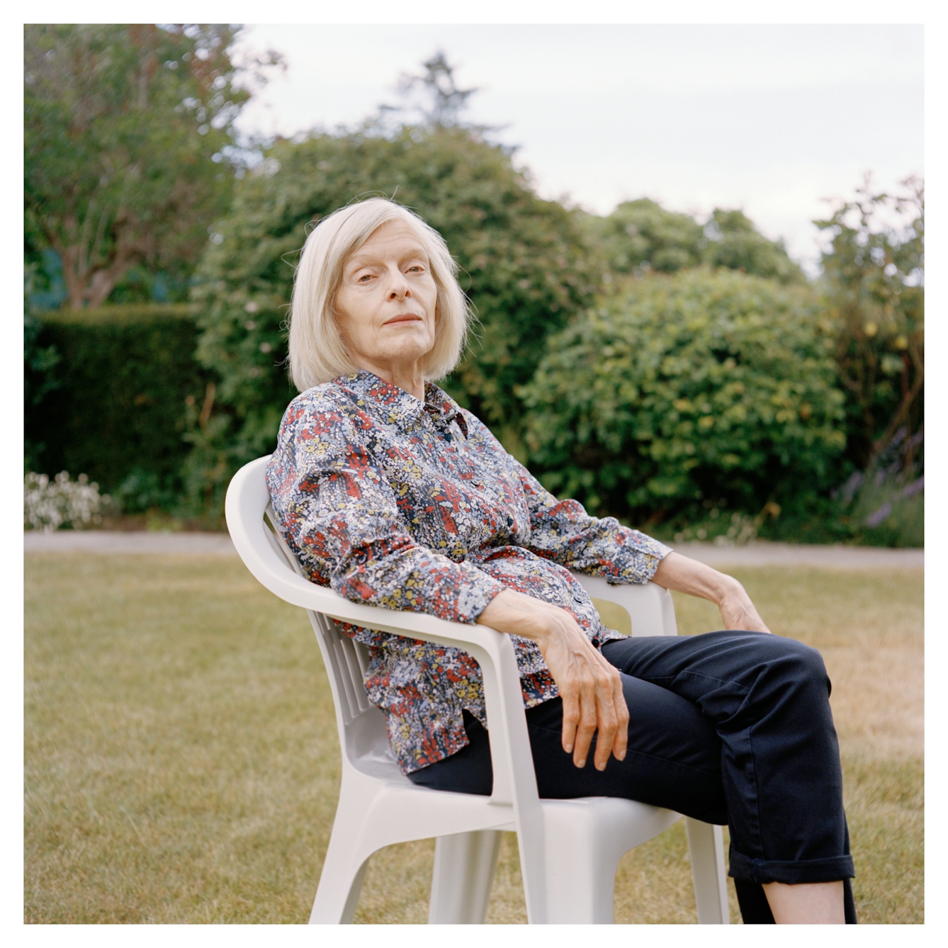 Portrait photograph of Margaret, an older woman with grey shoulder length hair. She is sitting in a white plastic garden chair in a garden.

Her body is sat sideways, with her head turned to face towards the camera. Her arms are resting on the chair arms. 

She is wearing a floral patterned shirt and black trousers. She is looking directly at the camera, with a neutral expression.