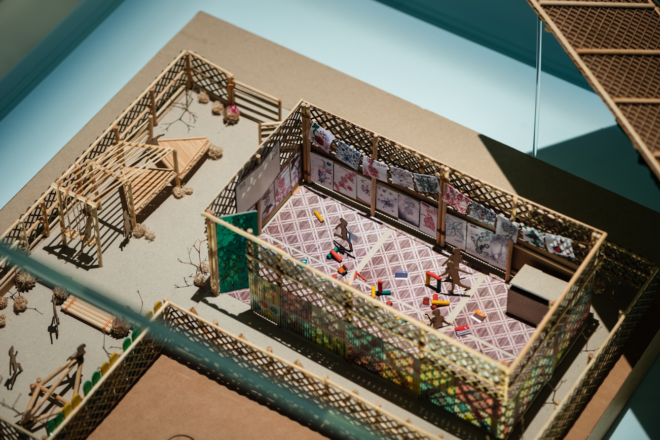 Photograph of a model of a buildings and outside spaces made in wood. Inside one of the structures are small figures representing children who are in the process of playing with colourful blocks.