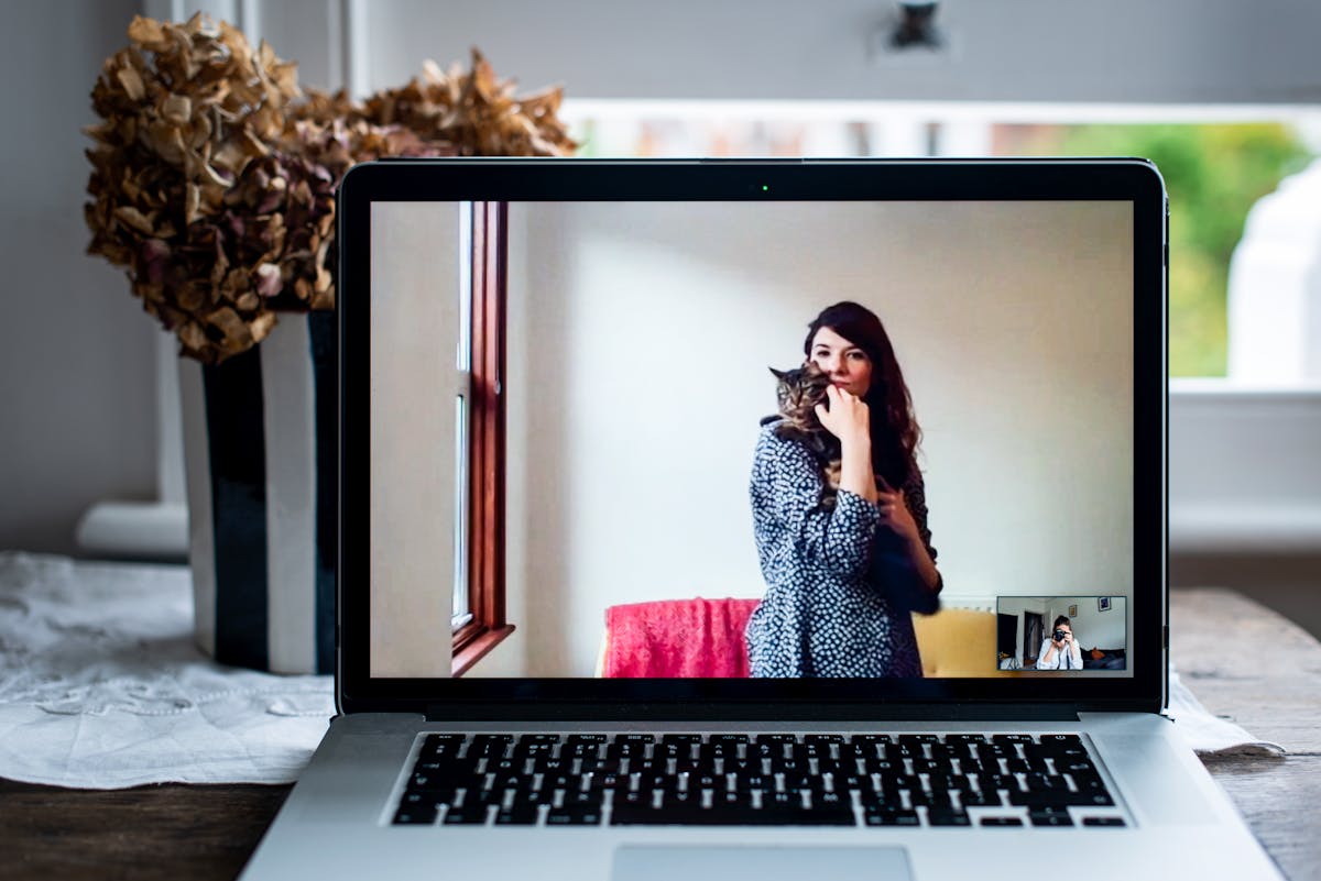 Photograph of an open laptop within a domestic scene. Most of the image is taken up with the screen, with part of the keyboard and trackpad visible. On the screen is a video call showing a woman standing in her living room, holding a cat in her arms, both are looking to camera. In the bottom right corner of the screen the photographer can be seen in a small floating window, camera to her eye, in the process of taking the picture.