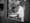 Still from black and white film featuring a doctor wearing a lab coat working. He is framed by the window of a door which has a label on it saying 'Hot lab', 'Radioactive Area'