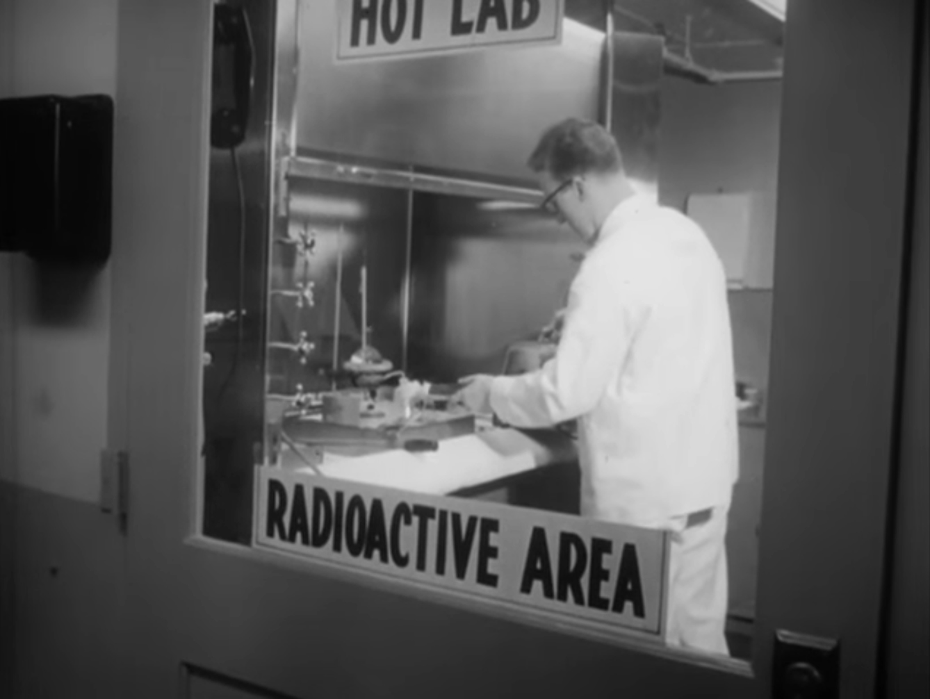 Still from black and white film featuring a doctor wearing a lab coat working. He is framed by the window of a door which has a label on it saying 'Hot lab', 'Radioactive Area'