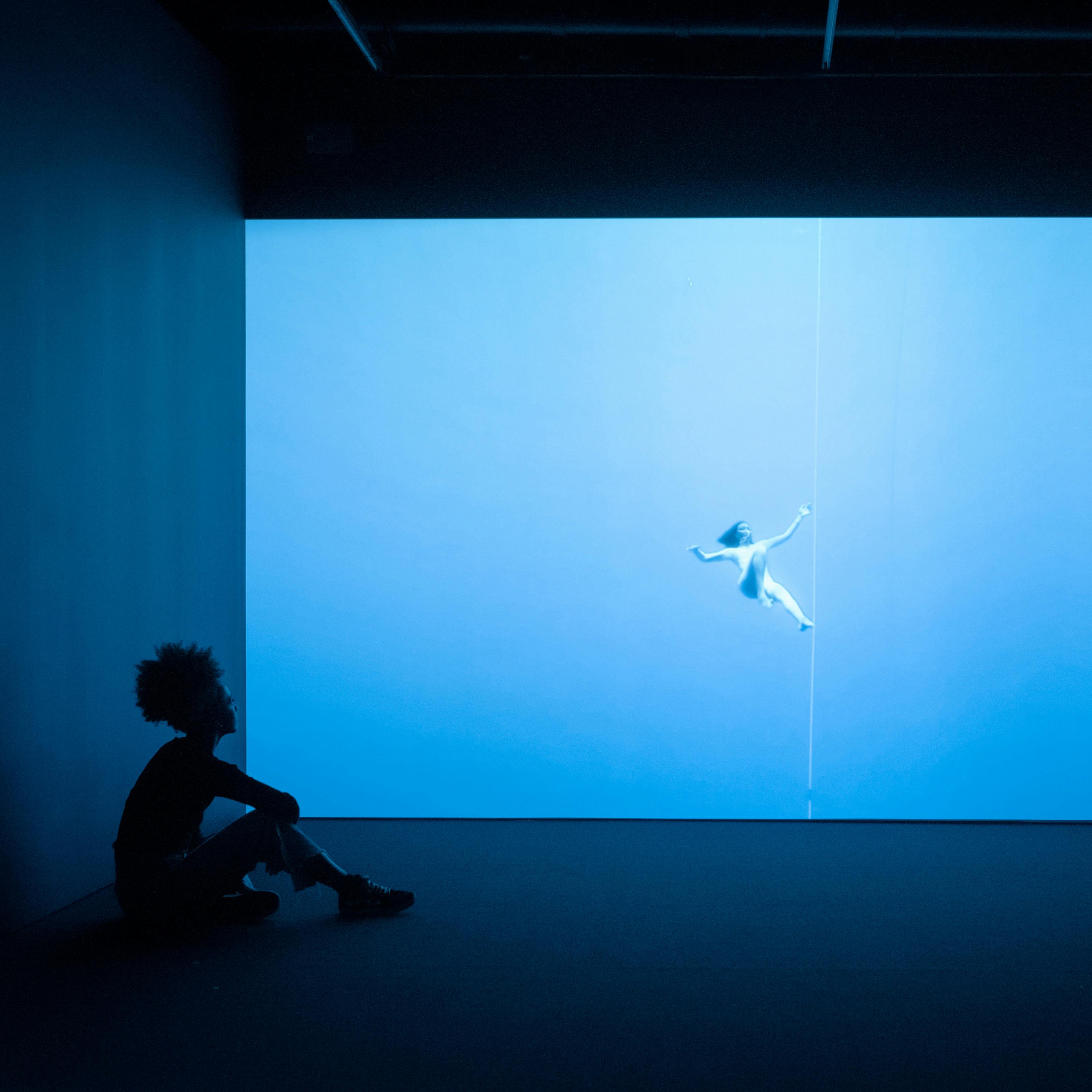 Photograph of a visitor exploring a gallery installation. She is sitting in the dark on the floor, in front of a large projection of a figure floating underwater. The room has a blue cast.