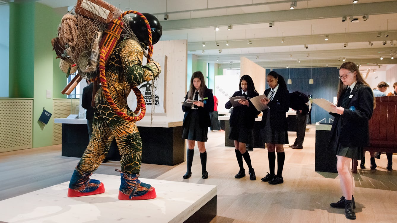 School students holding clipboards standing in a gallery space around a life-sized artwork of a figure resembling an astronaut 