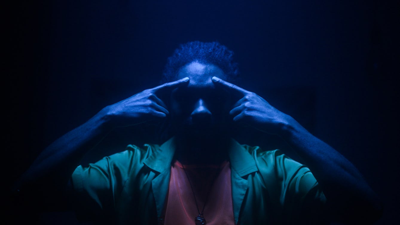 Film still of a man in a dark space, lit with a single blue light from directly above his head, casting much of his face into shadow. He is only visible from the chest up. He is wearing a green shirt, a light red t-shirt and a chain necklace. Both his arms are raise with both his index fingers pointing to his forehead, just above his eyebrows.